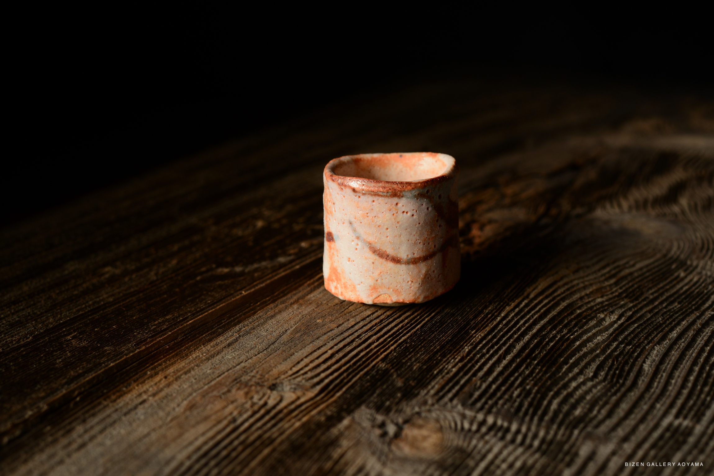A handcrafted Shino Guinomi sake cup, featuring a rustic design with earthy tones, resting on a textured wooden surface.