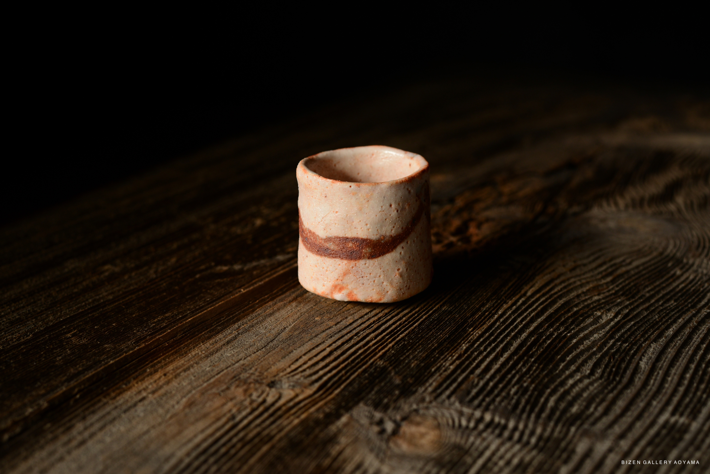 A close-up image of a Shino Guinomi, a traditional Japanese sake cup, featuring a light-colored ceramic body with a brown swirl pattern, sitting on a textured wooden surface.