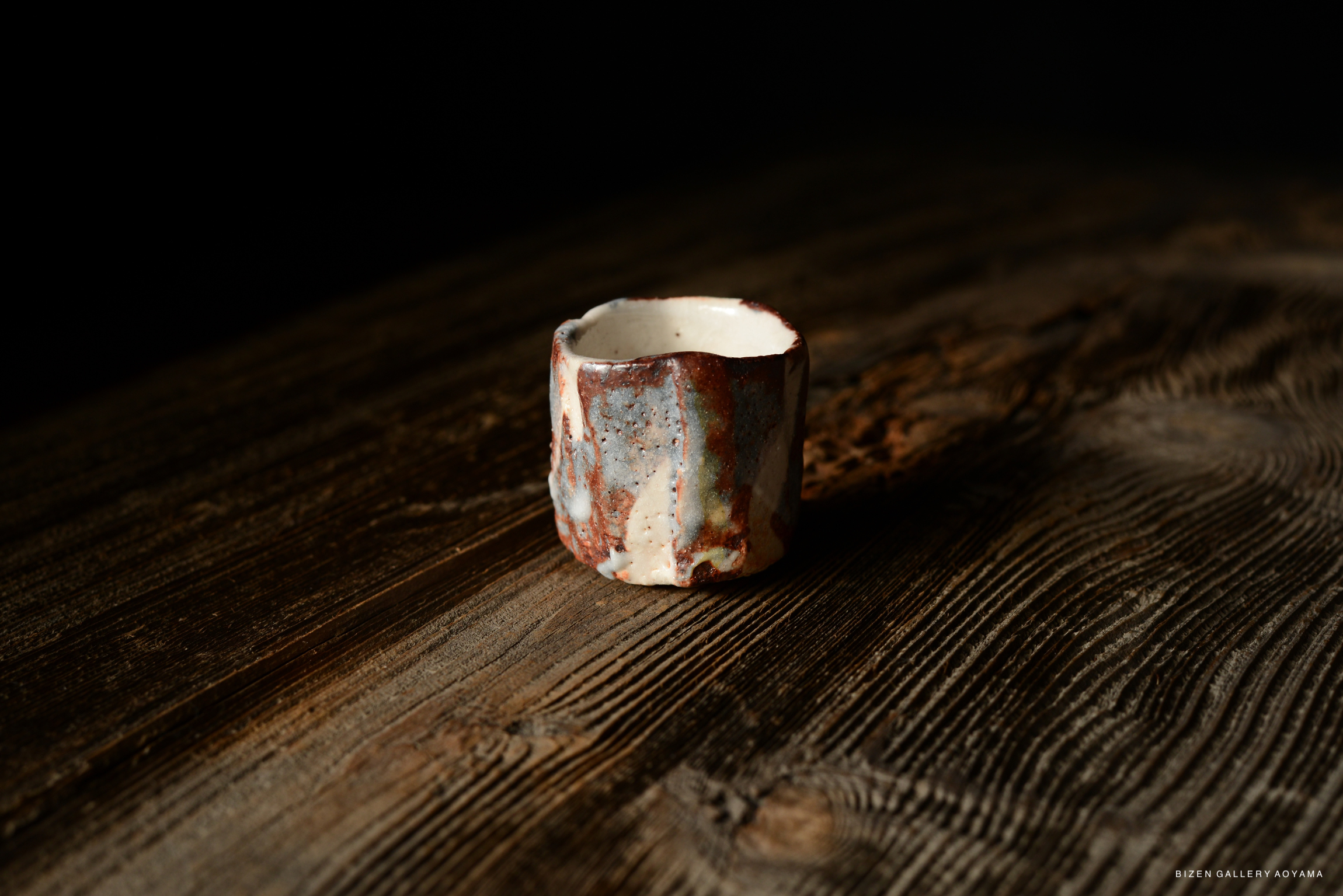 A small ceramic cup with a textured surface and a mix of earthy colors, placed on a rustic wooden table.