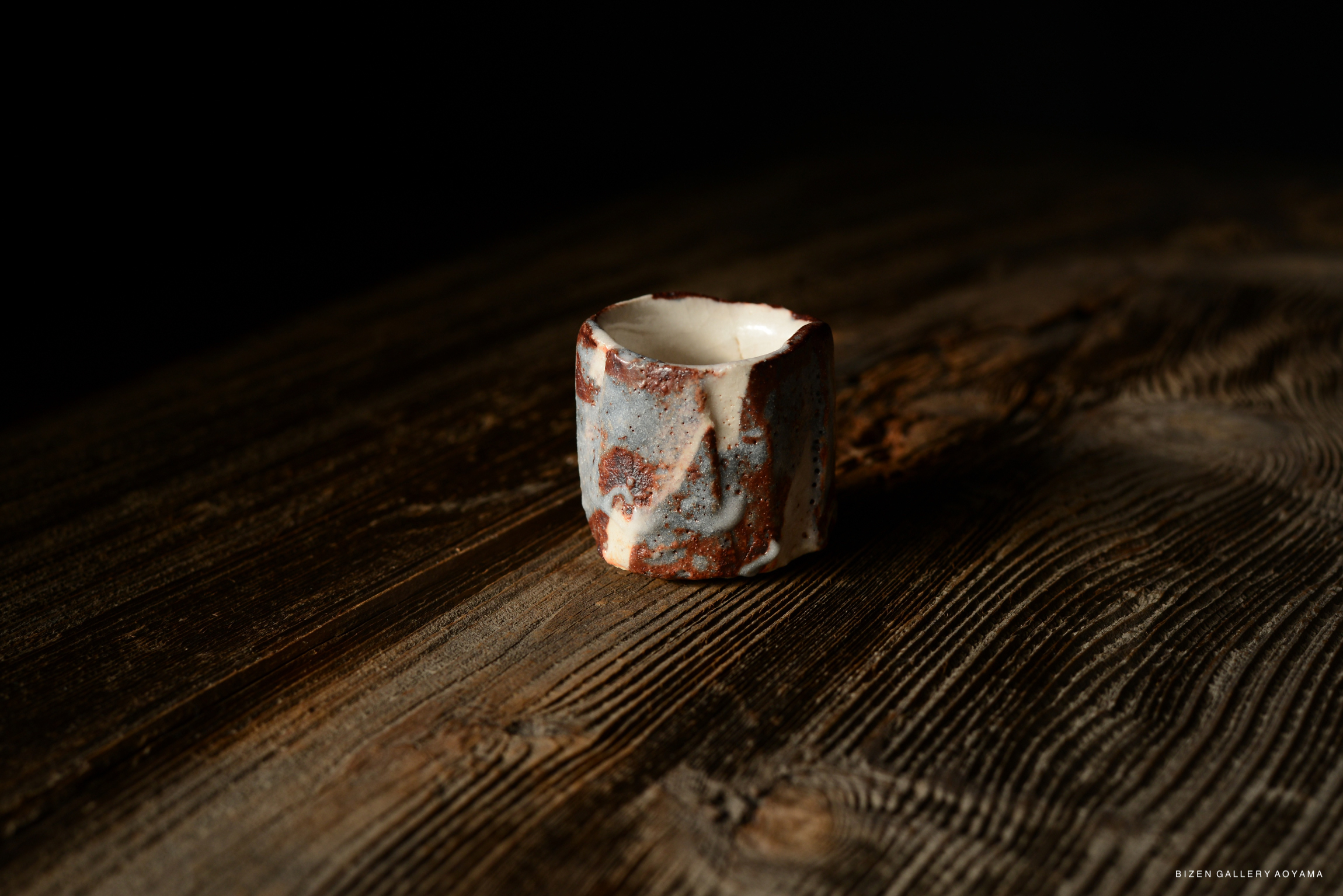 A Shino guinomi (sake cup) with a textured surface, displaying shades of blue and brown, resting on a wooden table.
