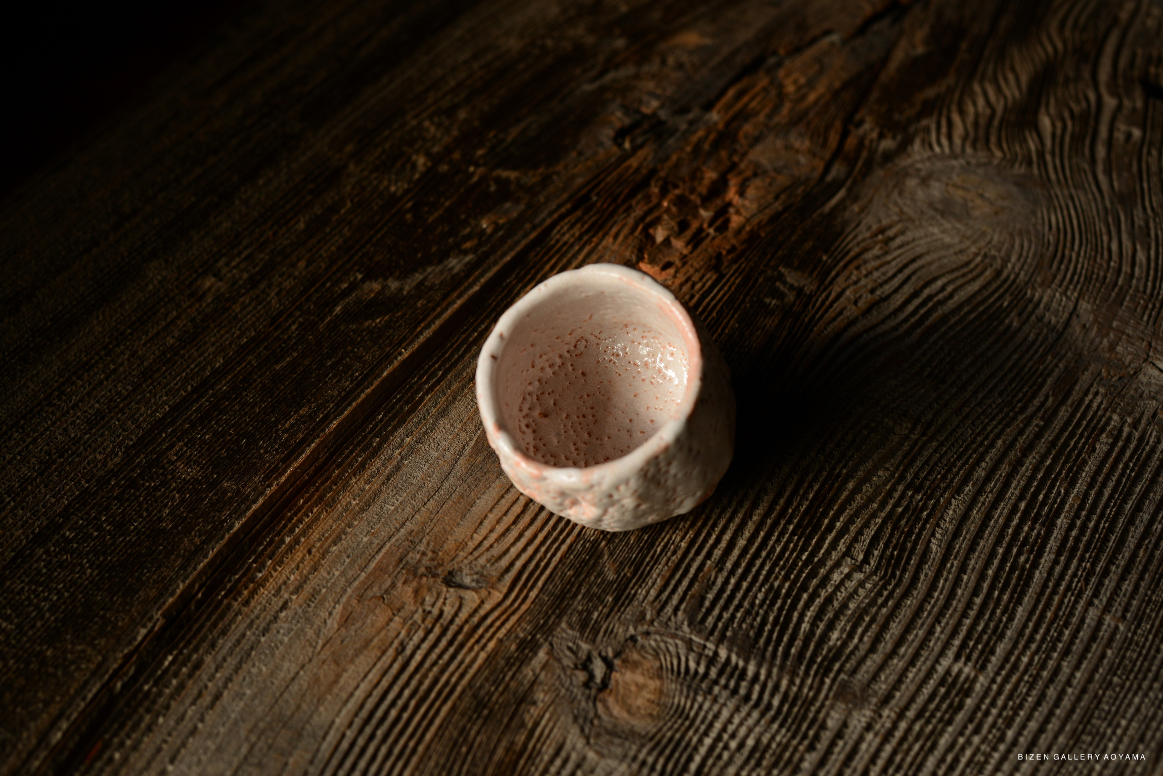 A close-up view of a small, handcrafted Shino sake cup resting on a rustic wooden surface.