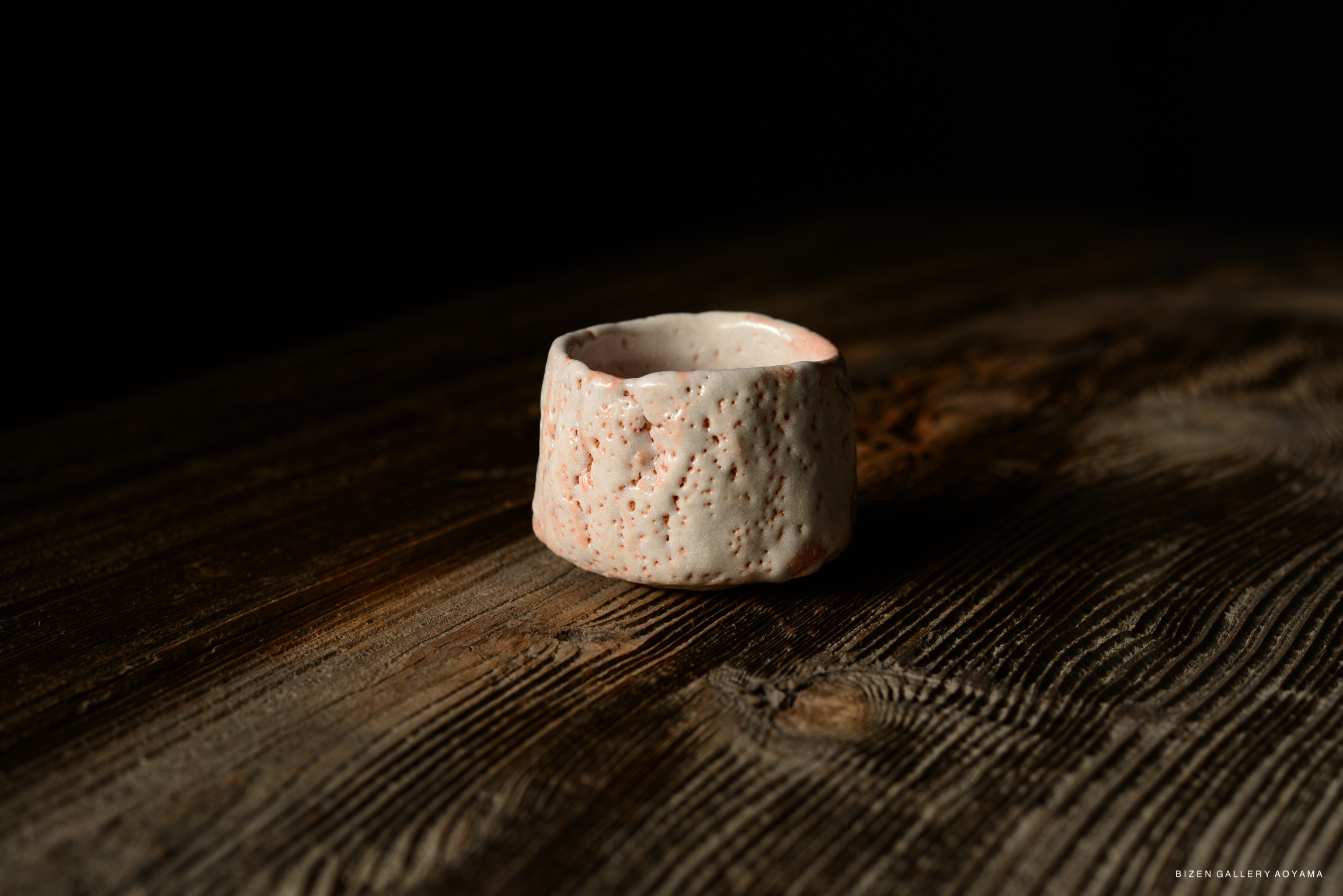 A close-up of a Shino Guinomi sake cup with a textured white and light pink surface, placed on a wooden table.