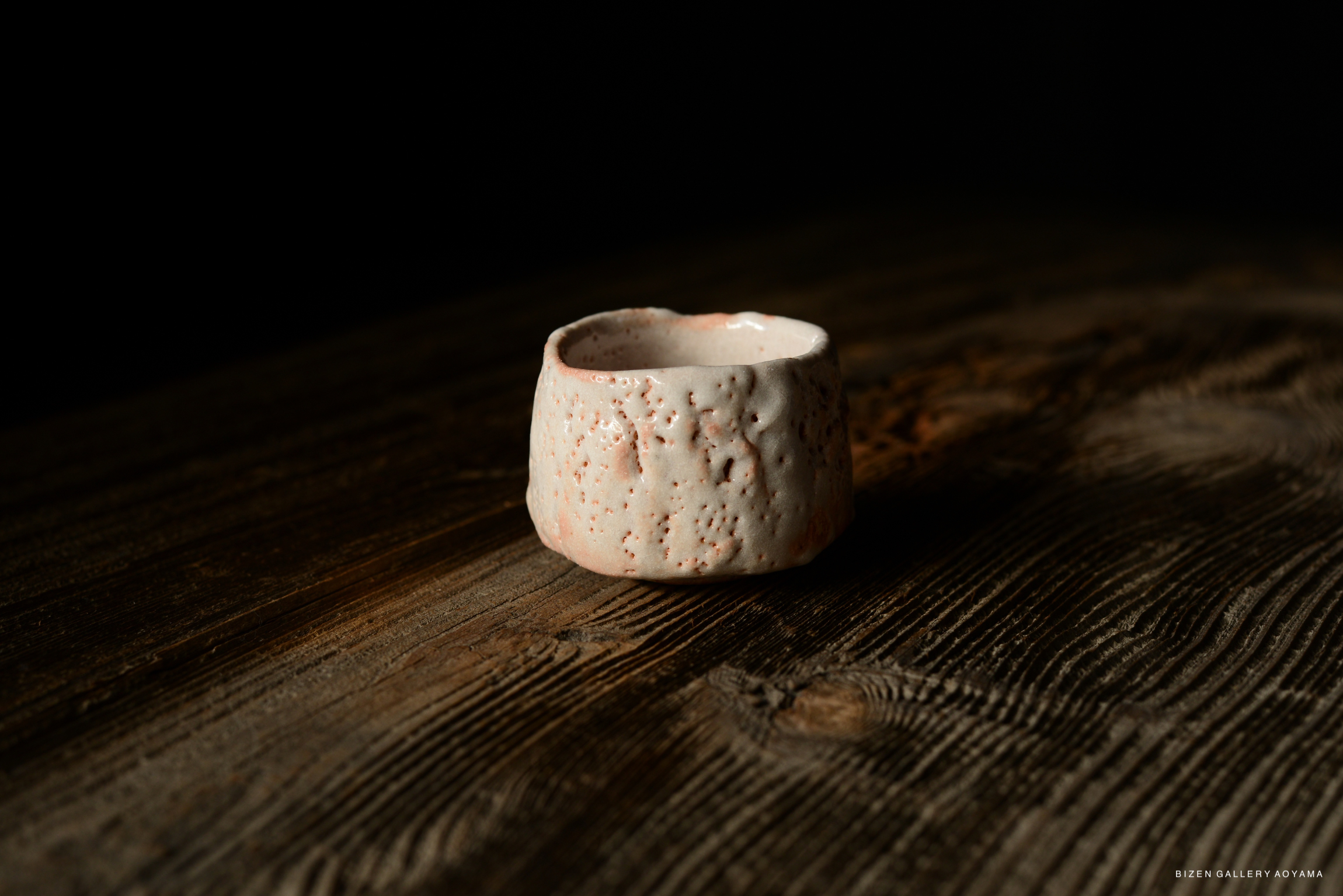 A close-up of a Shino Guinomi sake cup with a textured surface and a light pinkish hue, placed on a rustic wooden table.
