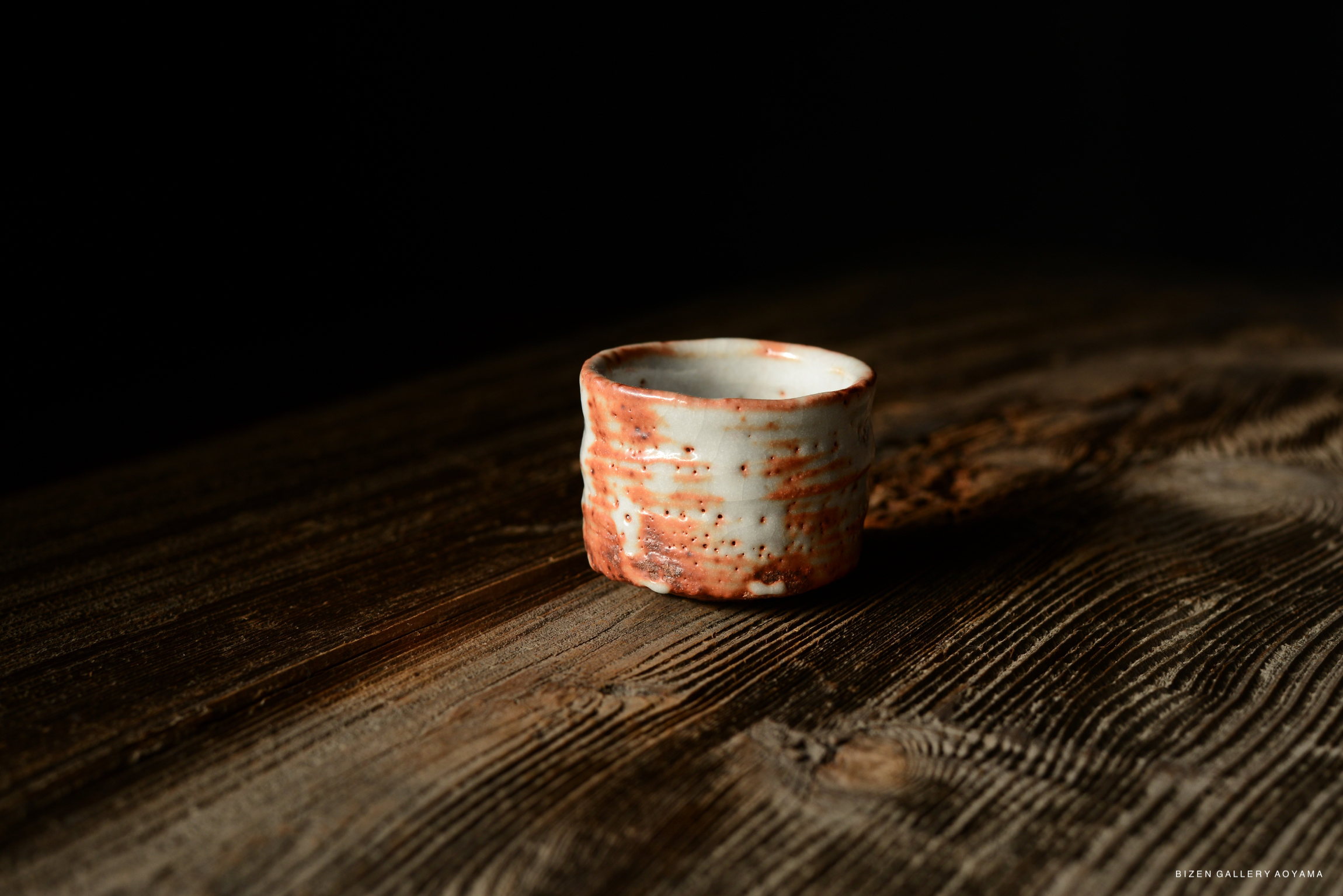 A small ceramic cup with a textured surface and reddish accents, resting on a wooden table with a dark background.
