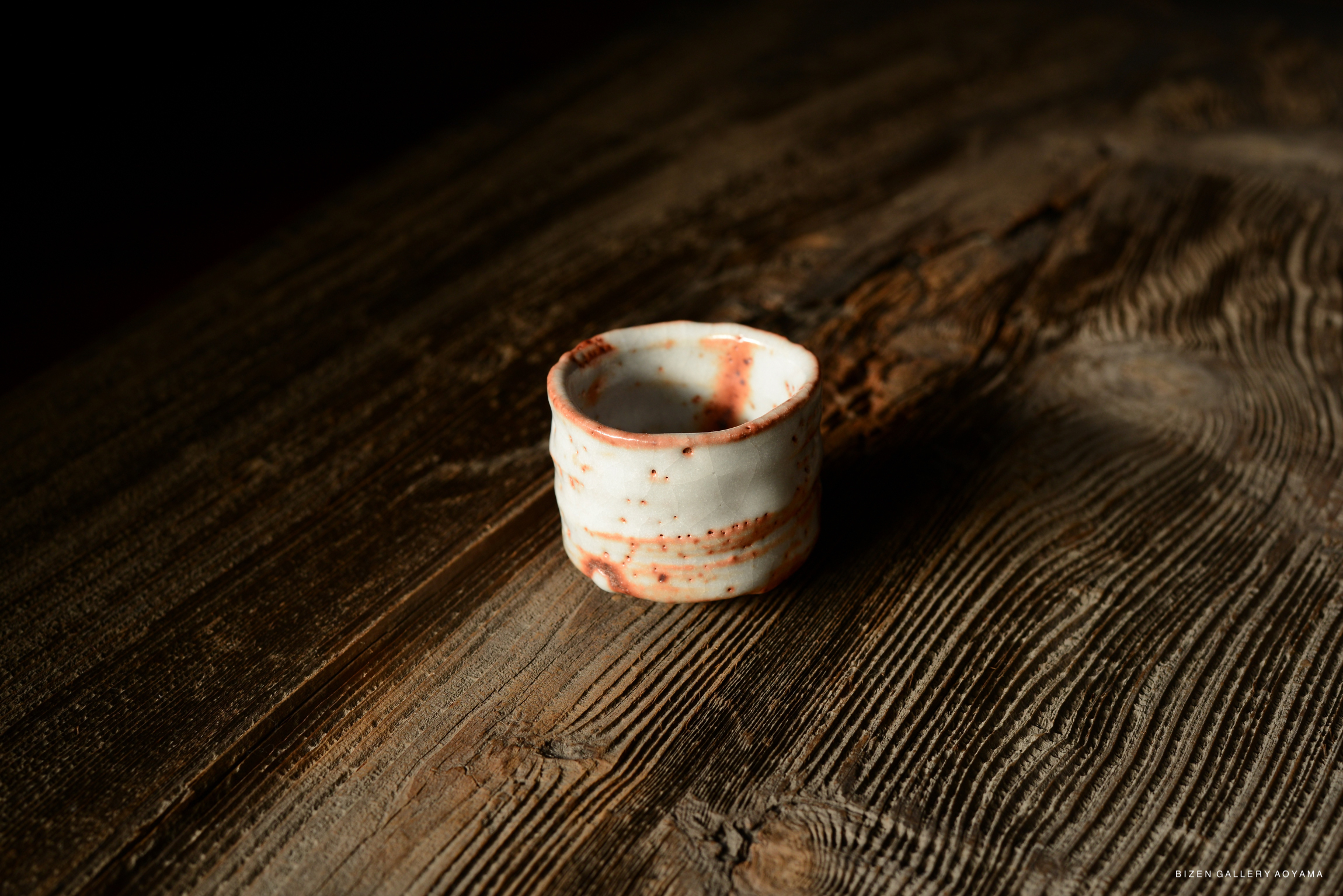 A small, handmade ceramic cup with a white and reddish-brown speckled design, placed on a textured wooden surface.