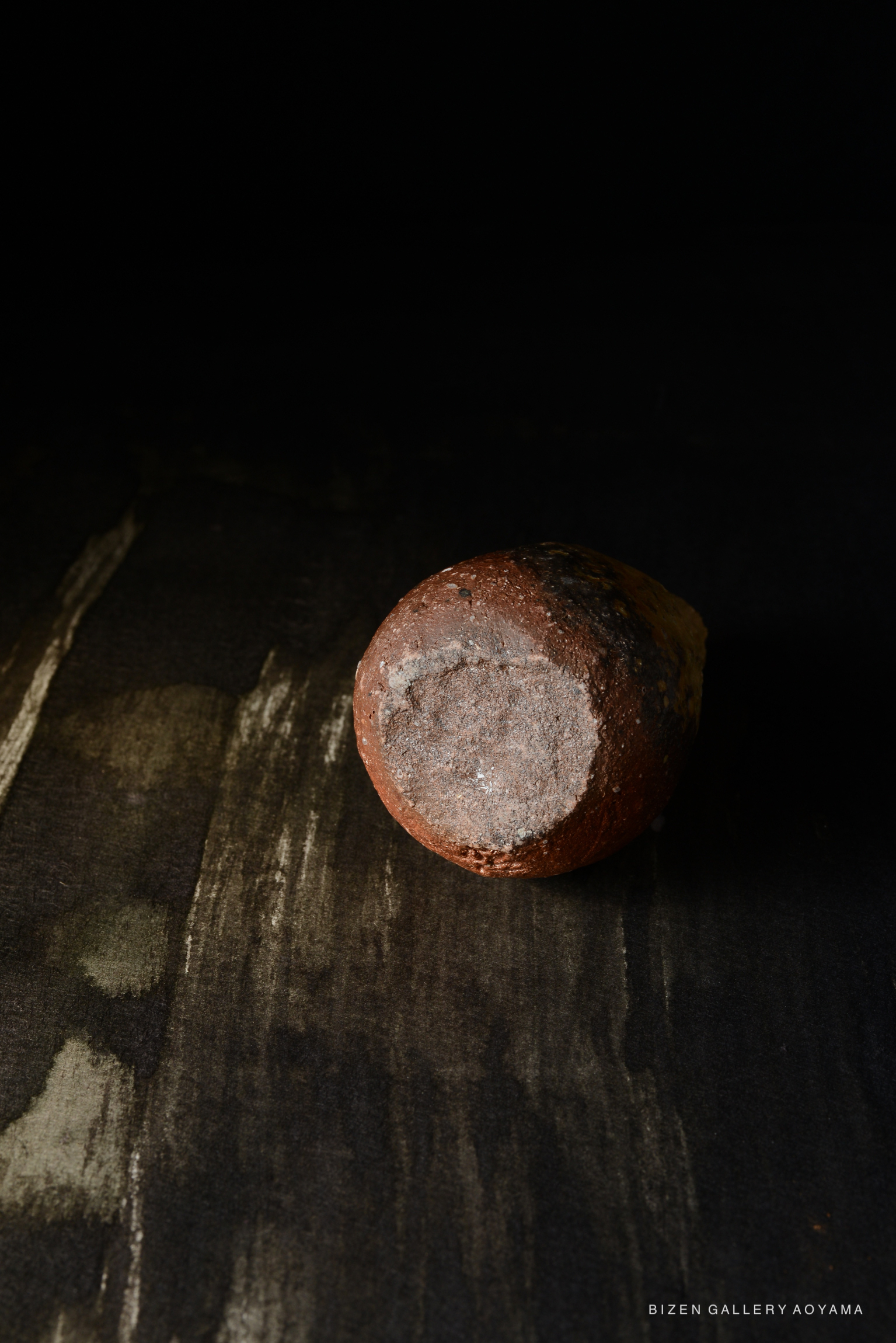 暗い背景に置かれた陶器の一部。表面は滑らかで、茶色と灰色の色合いが混在している。