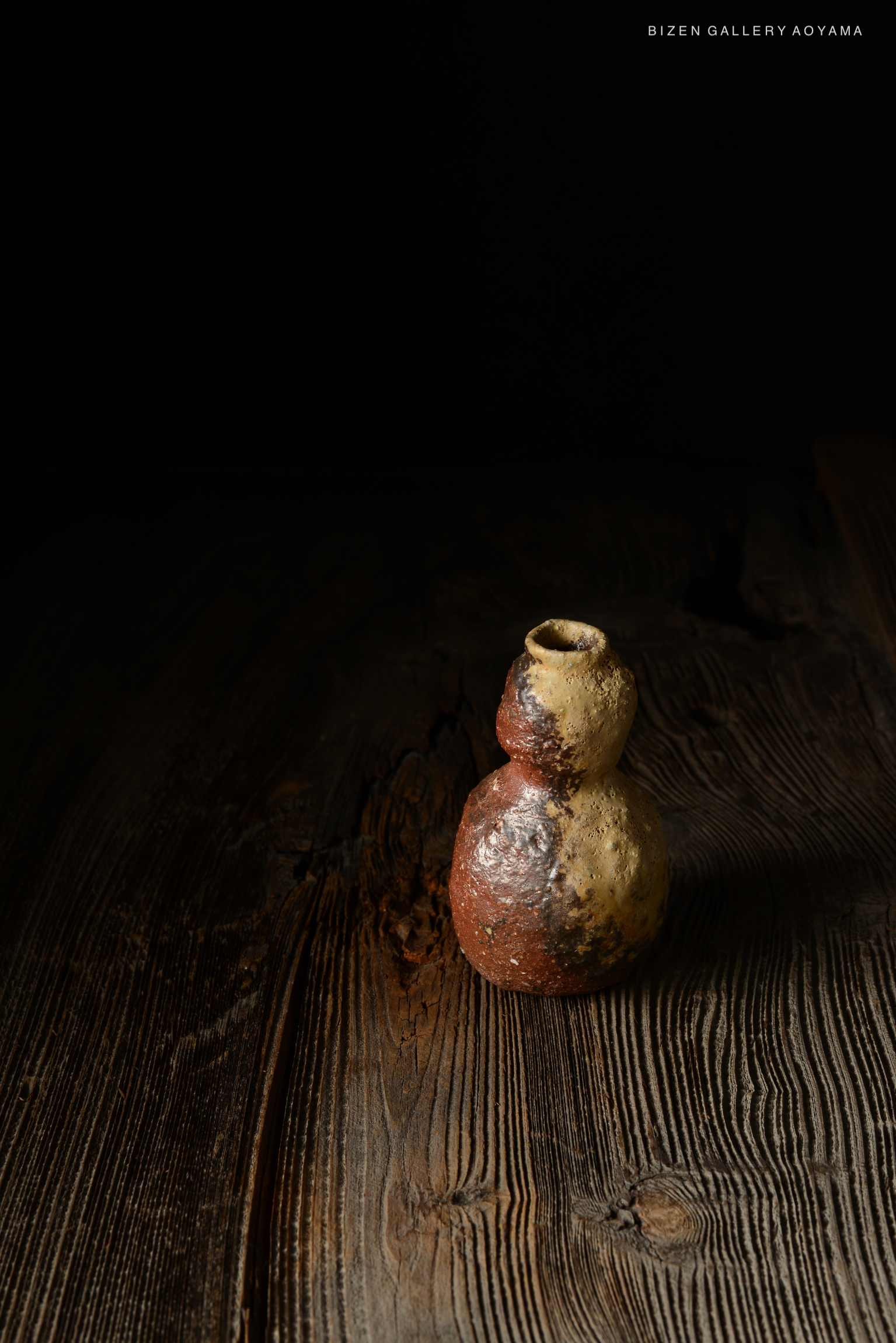 黒い背景の前に置かれた、独特な形状のビゼン焼きの陶器。木製のテーブルの上にあり、色合いは赤と黄色が混ざり合っている。