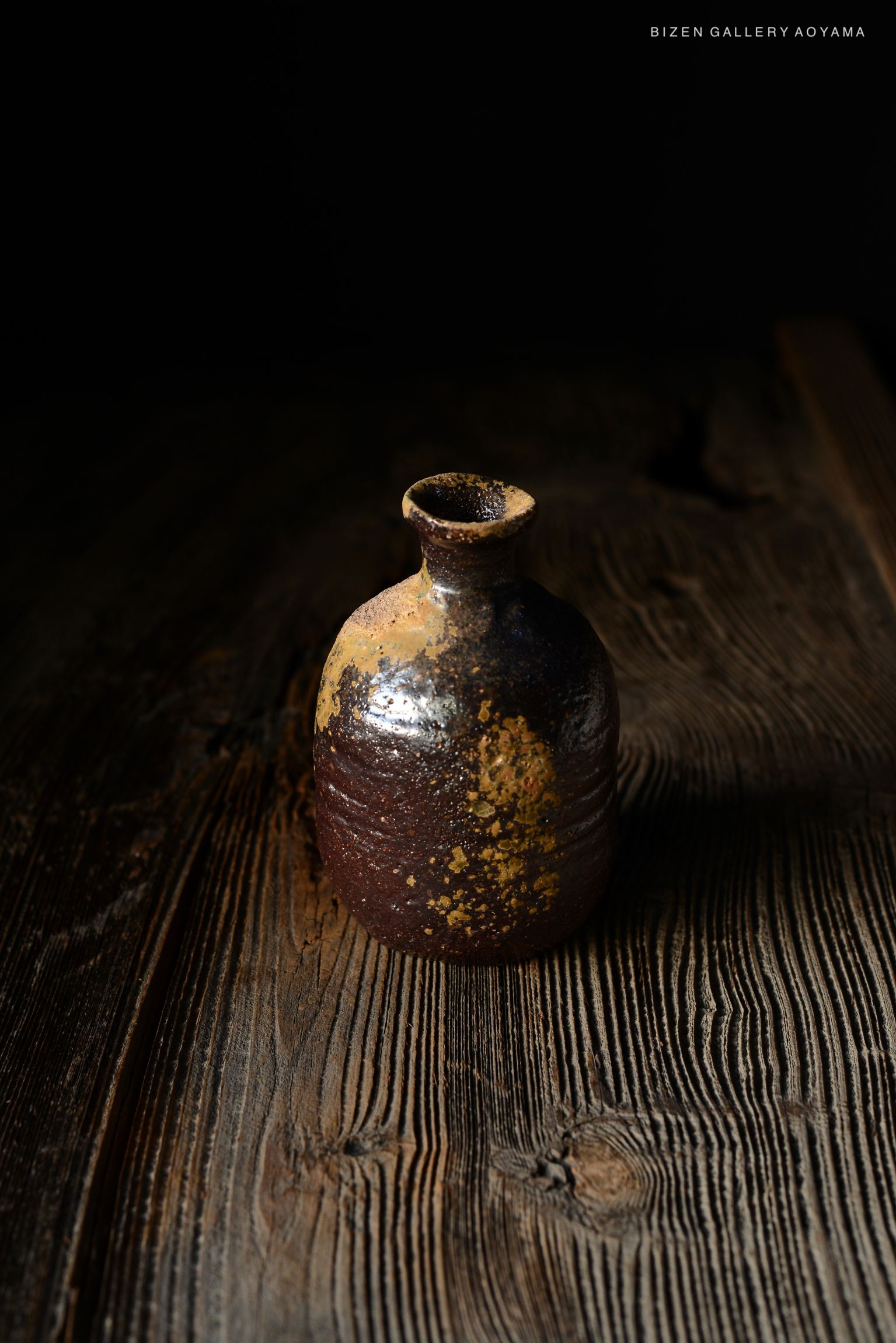 暗い背景の前に置かれた、古い日本の備前焼の瓶。木のテーブルの上にあり、独特の色合いと質感が特徴。