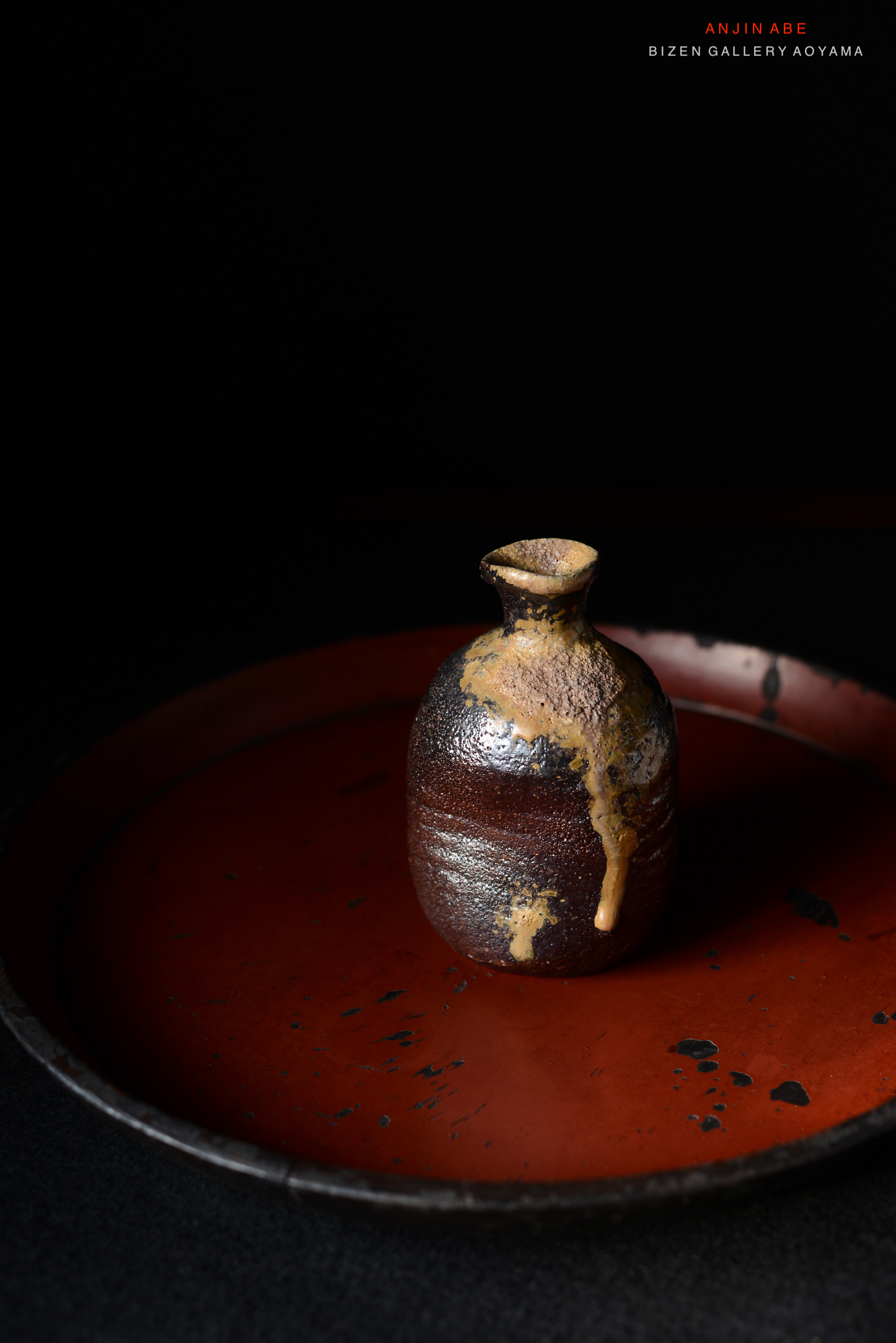 暗い背景の上に置かれた小さな備前焼の壺。赤い皿の上にあり、壺の表面には独特な釉薬の流れが見える。