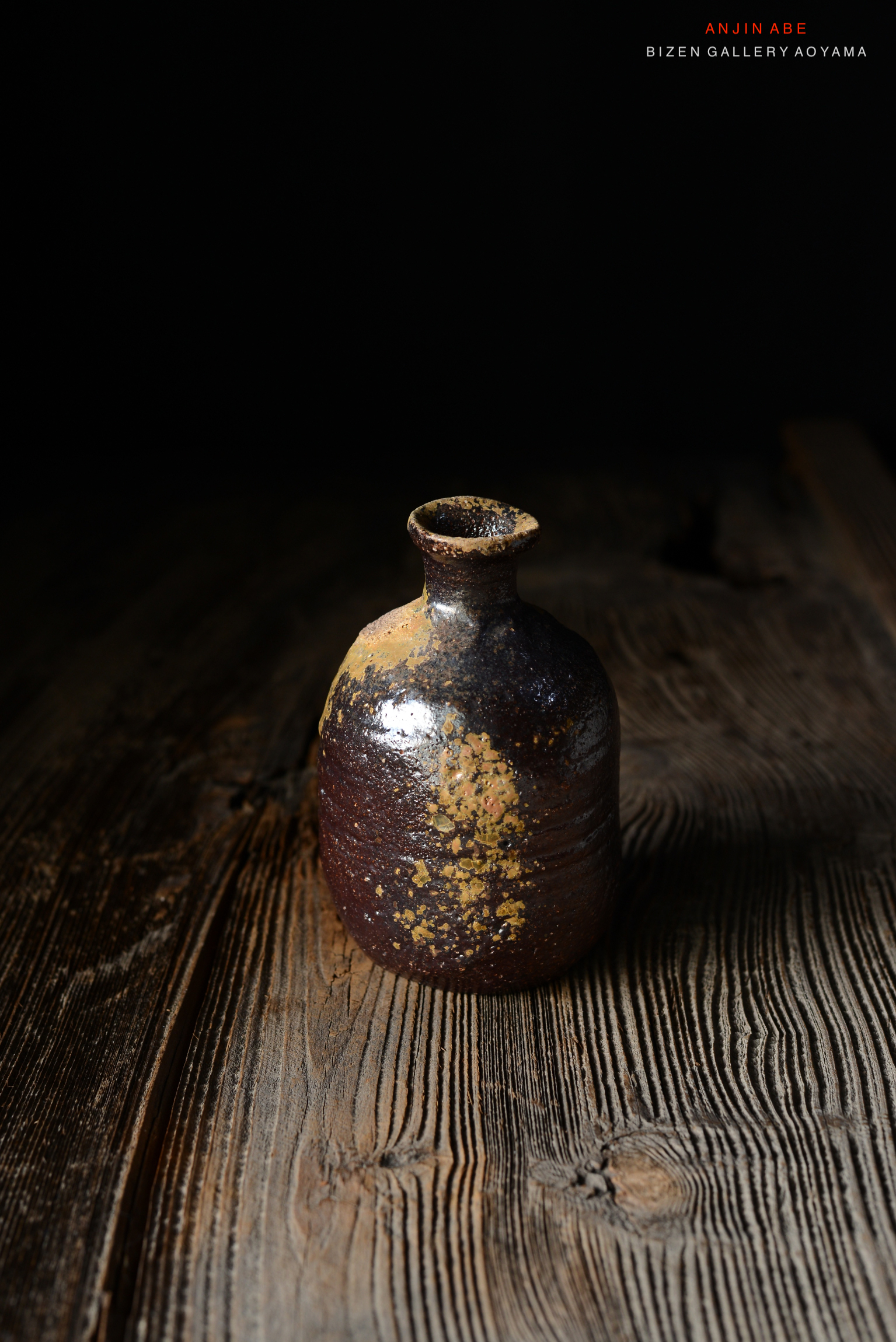 黒い背景に置かれた、ビゼン焼きの陶器の瓶。木製のテーブルの上にあり、独特の釉薬模様が見える。