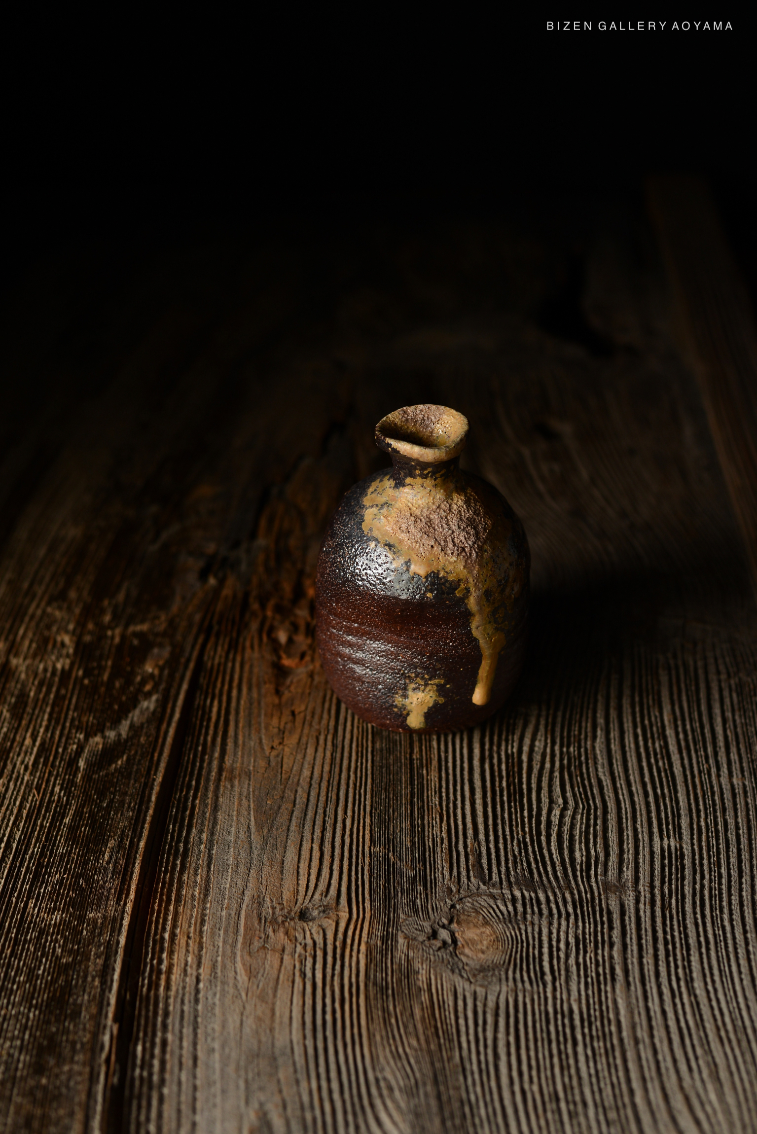 黒い背景の上に置かれたビゼン焼きの陶器。茶色と金色の釉薬がかかった独自の形状をした壺。木のテーブルの上に自然光で照らされている。