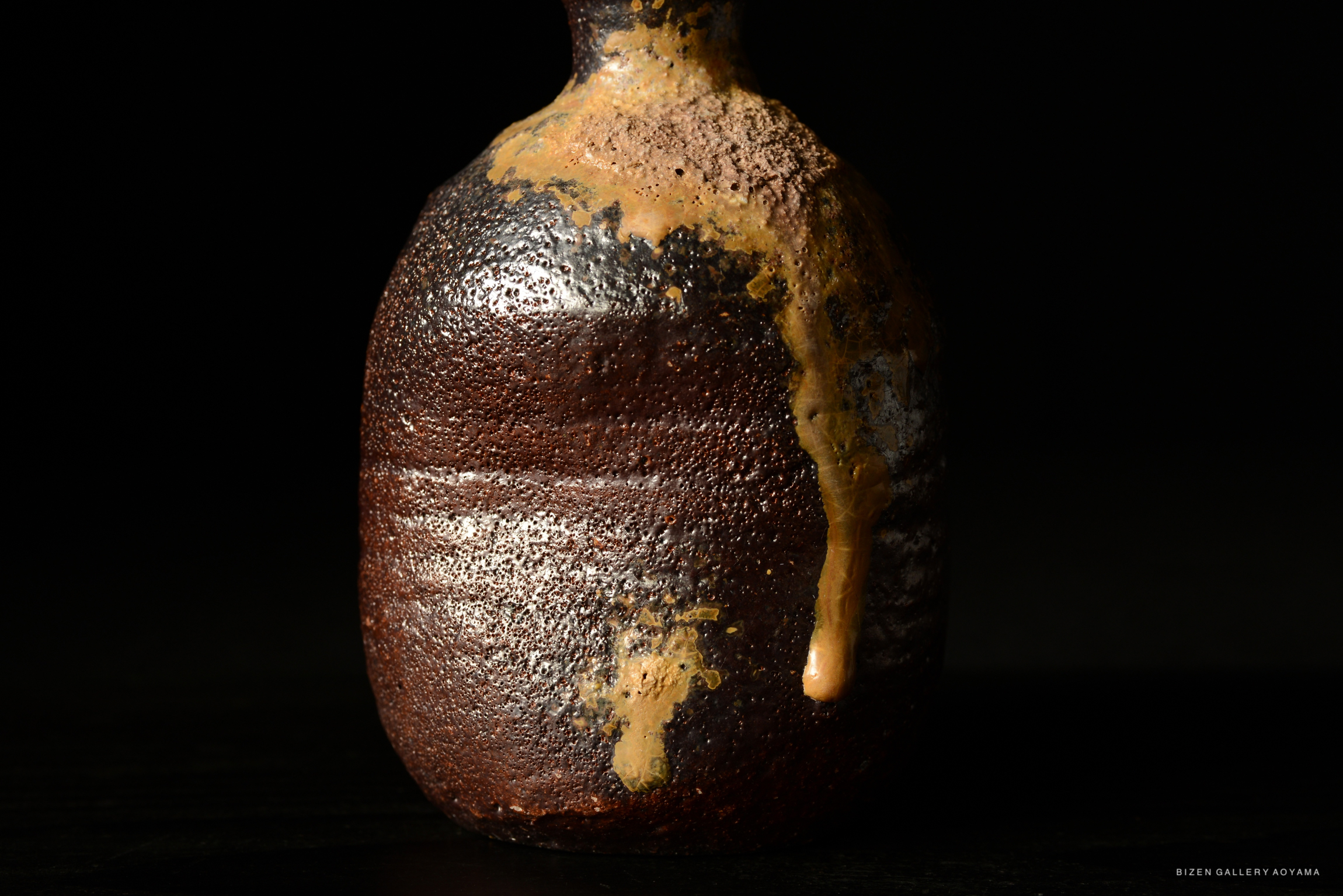 暗い背景に置かれた独特の形状の陶器の瓶。表面には焦げた部分と流れた釉薬が見える。