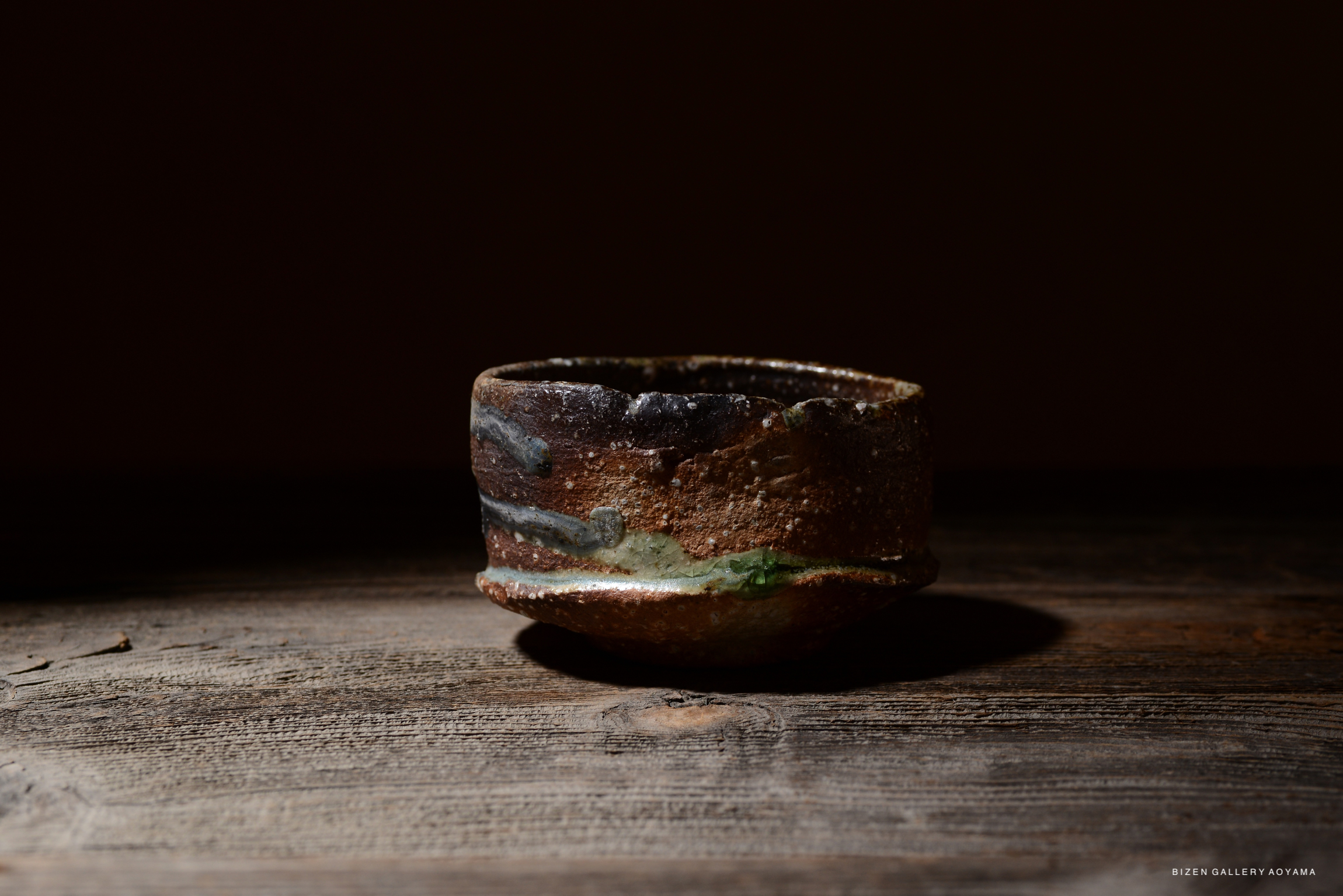 木のテーブルの上に置かれた、有機的な形状の茶碗。茶碗は茶色の釉薬と緑色のアクセントが施されている。暗い背景が特徴的で、茶碗が際立っている。