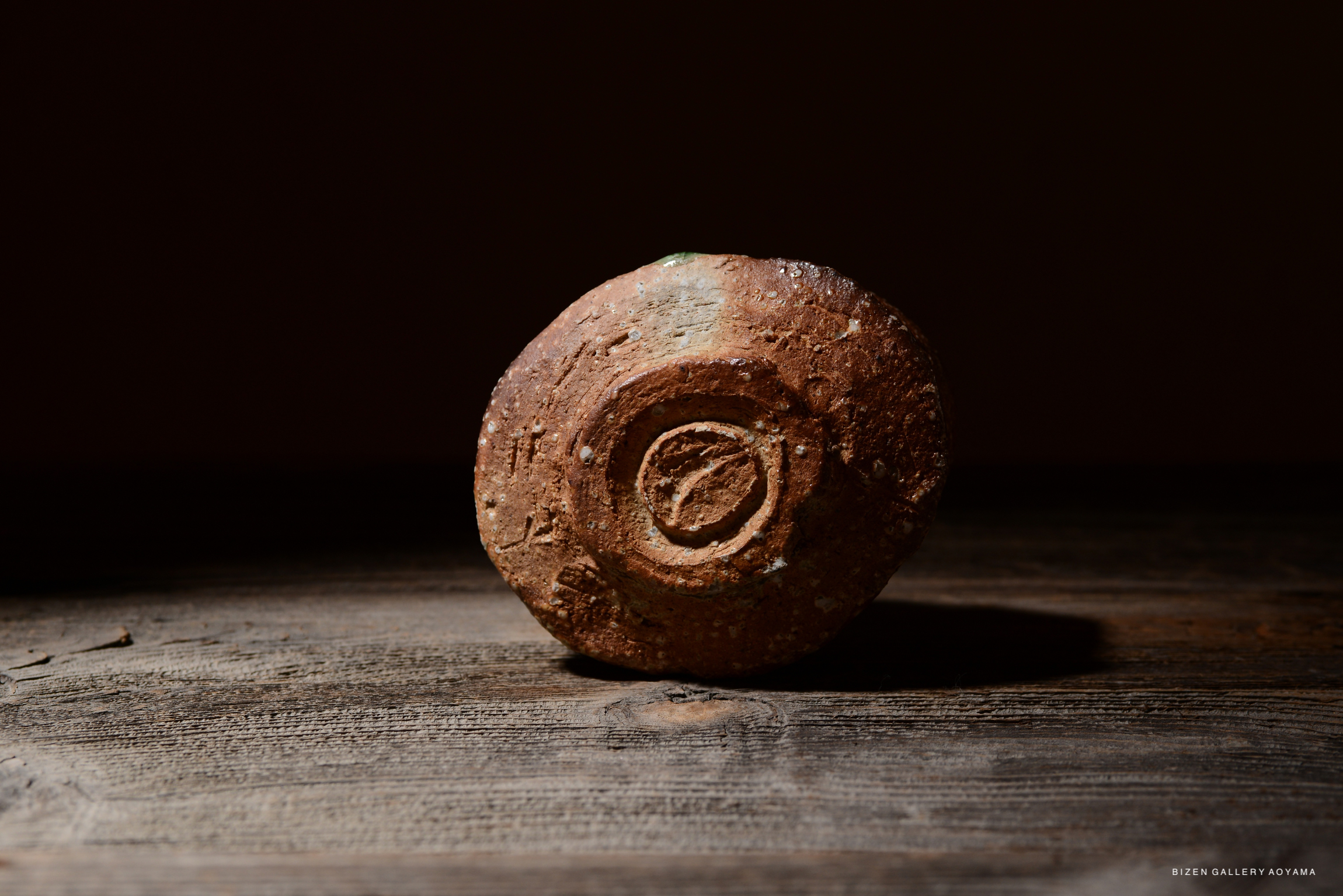 暗い背景の上に置かれた土製の壺のようなオブジェクト。表面には模様があり、自然な質感を持つ。木製のテーブルの上に配置されている。