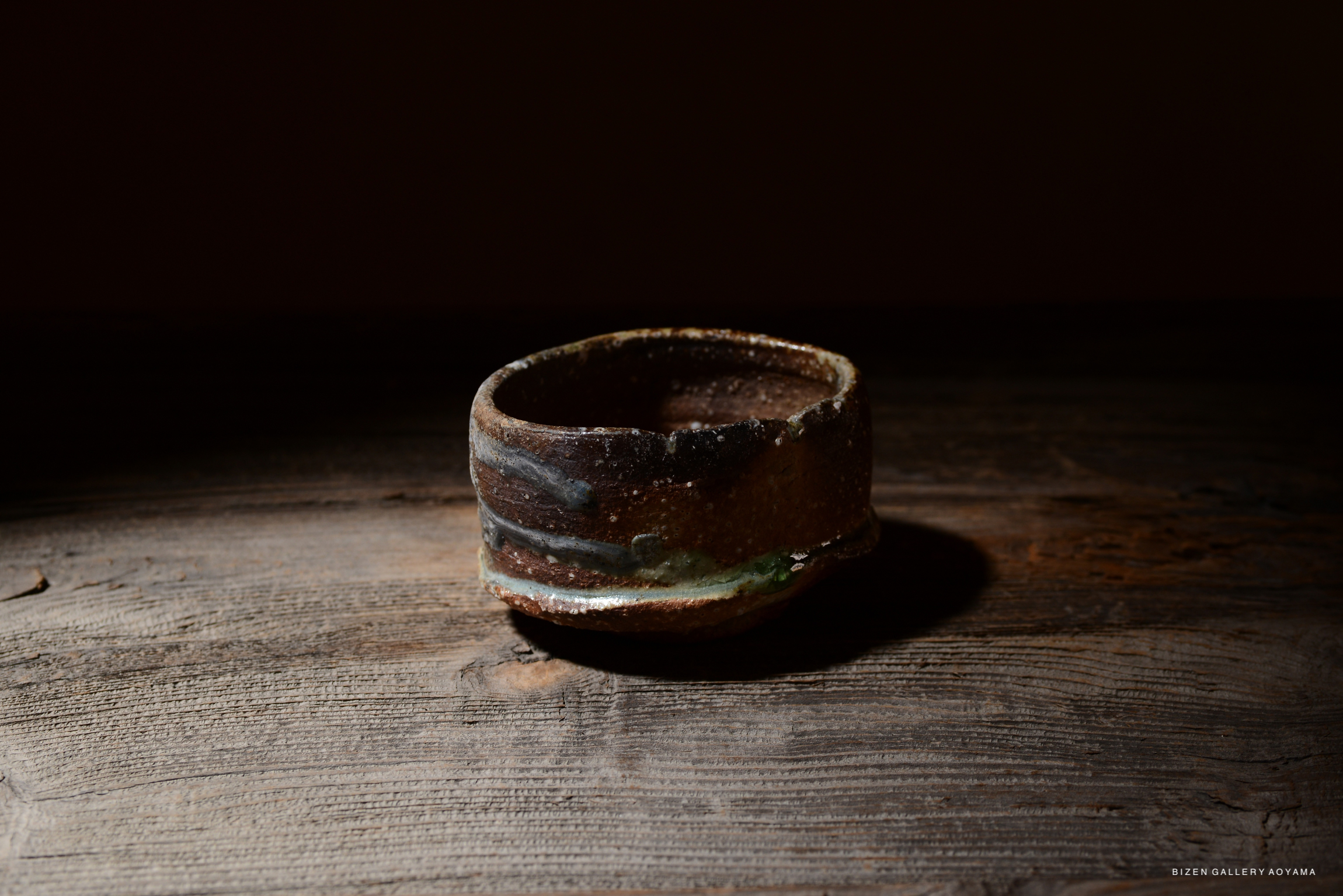 木のテーブルの上に置かれた、独特な陶器の器。茶色と緑の釉薬が施されており、土の質感が感じられる。