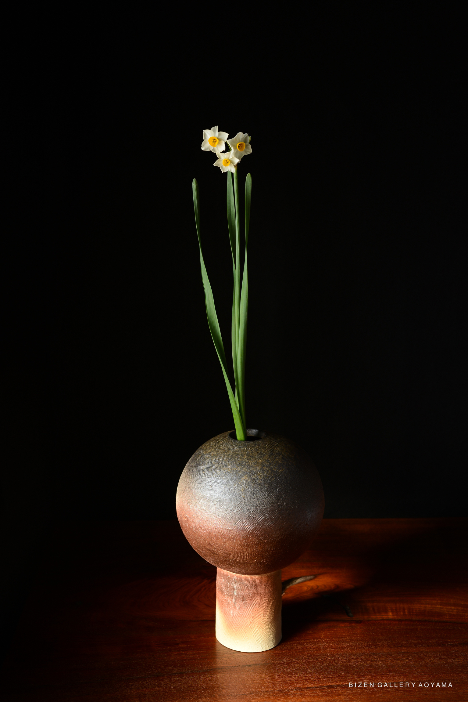 A close-up of a vase with a round top and a cylindrical base, displaying yellow-centered white flowers with long green leaves against a dark background.