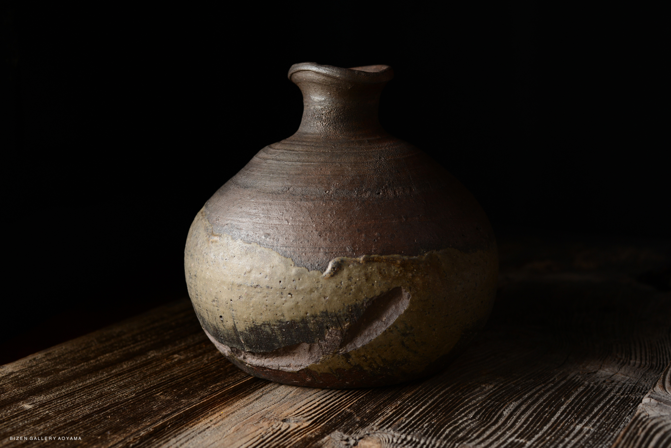 A close-up of a handcrafted ceramic jug with a smooth, rounded body and a narrow neck, resting on a textured wooden surface.