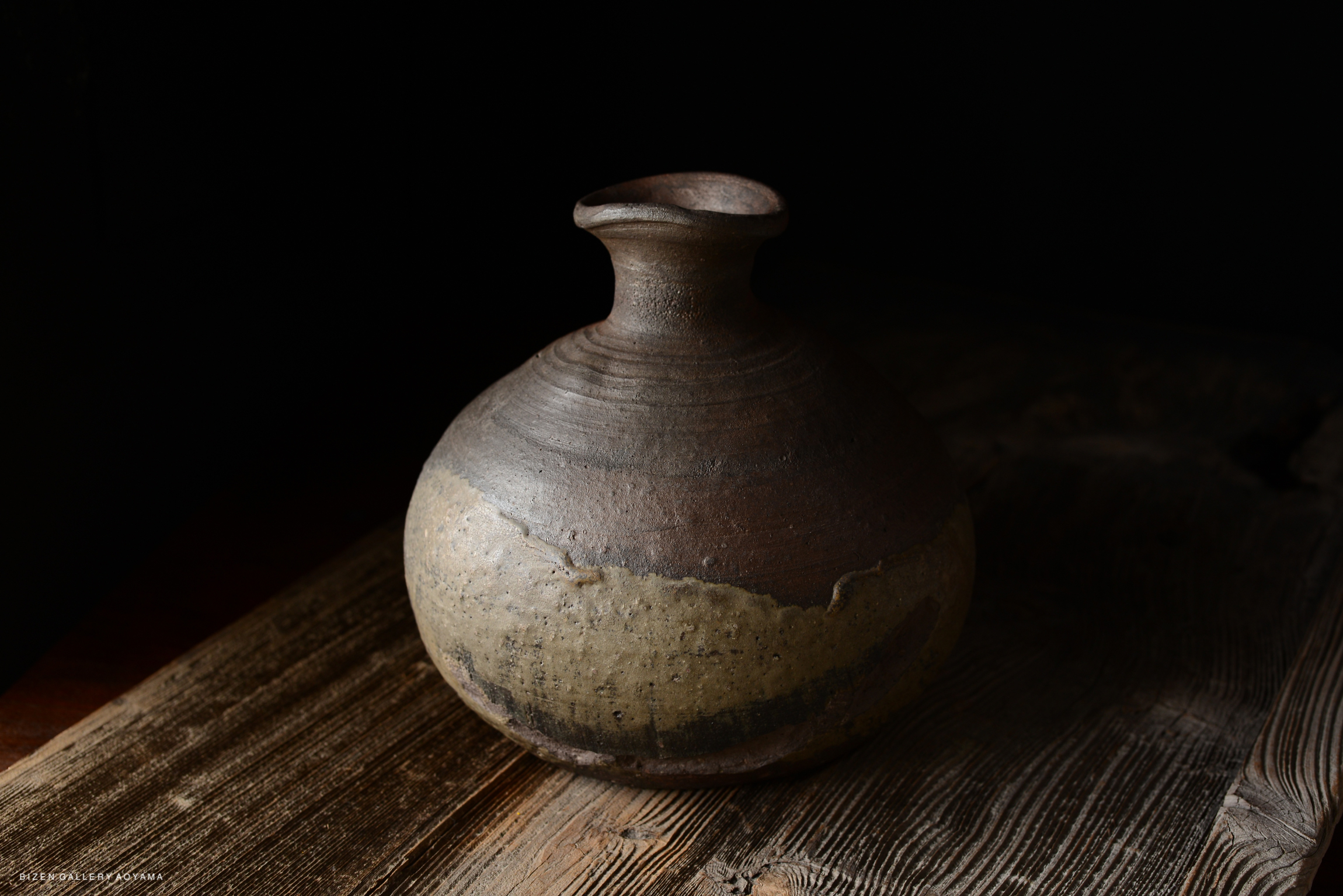 A round, textured clay pot with a narrow neck, placed on a wooden surface. The pot features a two-tone design with dark and light brown colors, against a dark background.