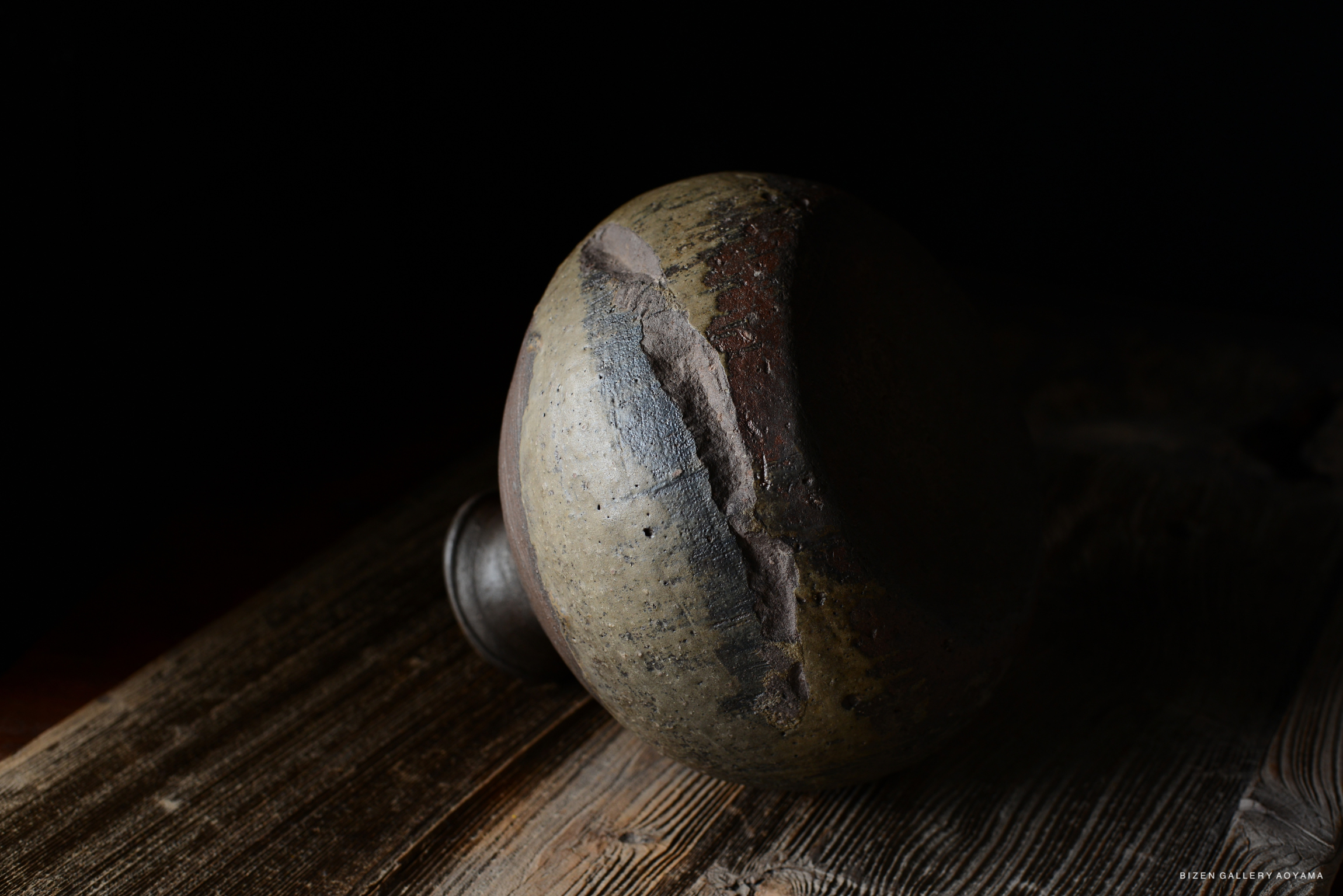 A close-up view of a traditional pottery piece with an earthy texture and various colors, resting on a wooden surface against a dark background.