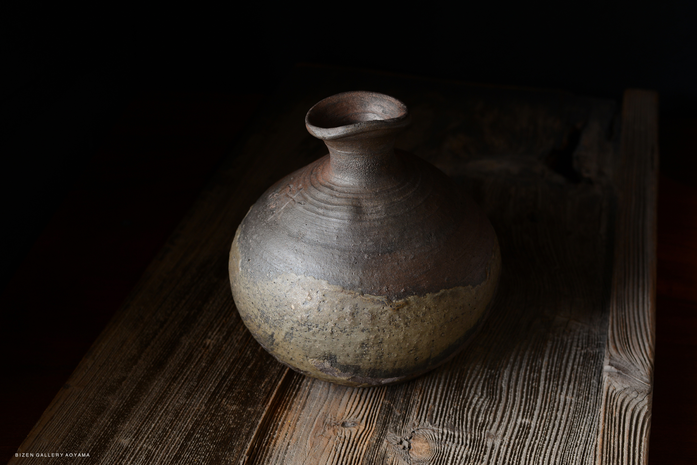 A handcrafted ceramic vase with a rounded body and a narrow neck, showcasing a natural clay texture and a mix of brown and beige glazes, placed on a rustic wooden surface.