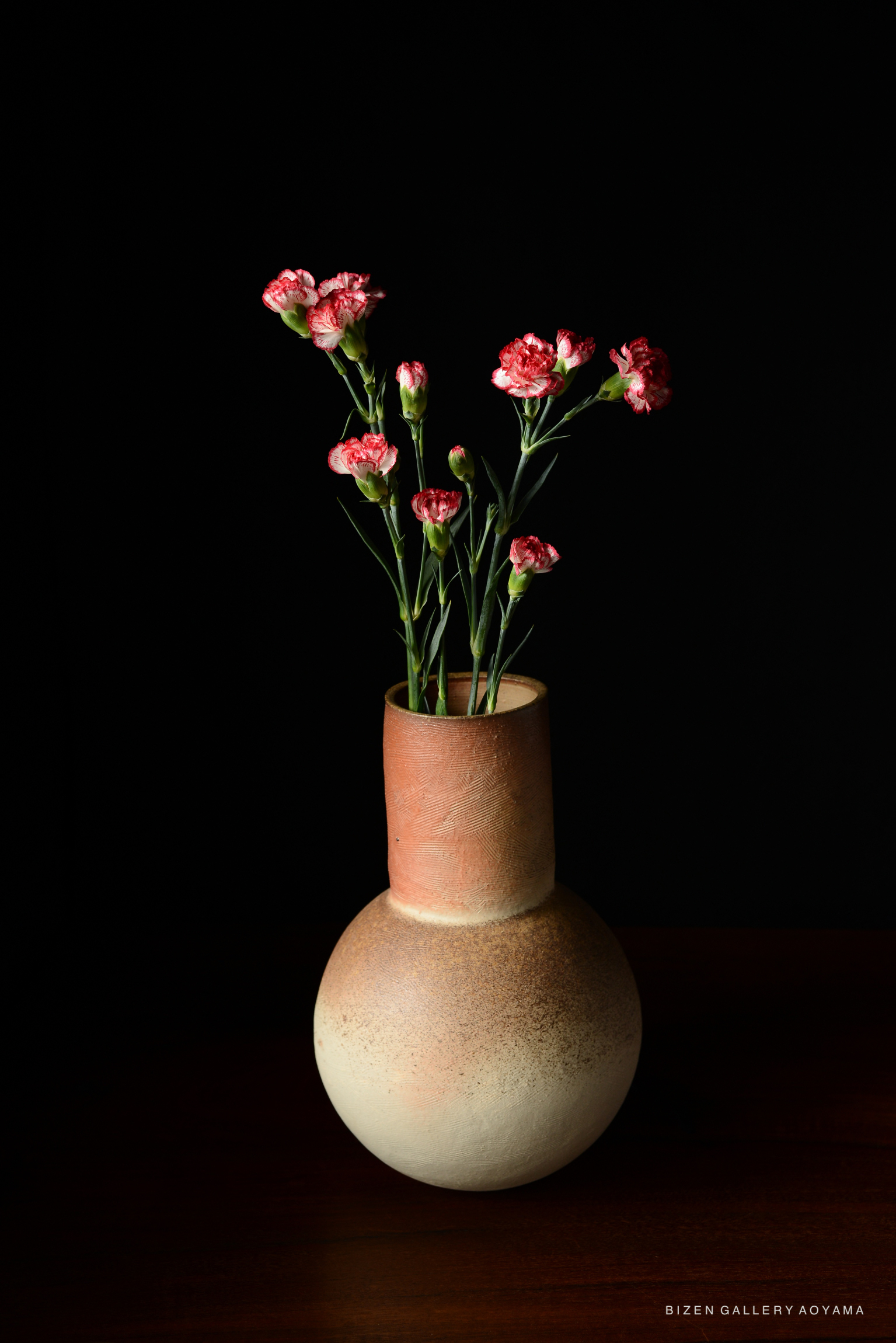 A simple ceramic vase with a round base and a narrow neck, filled with pink carnations, against a black background.