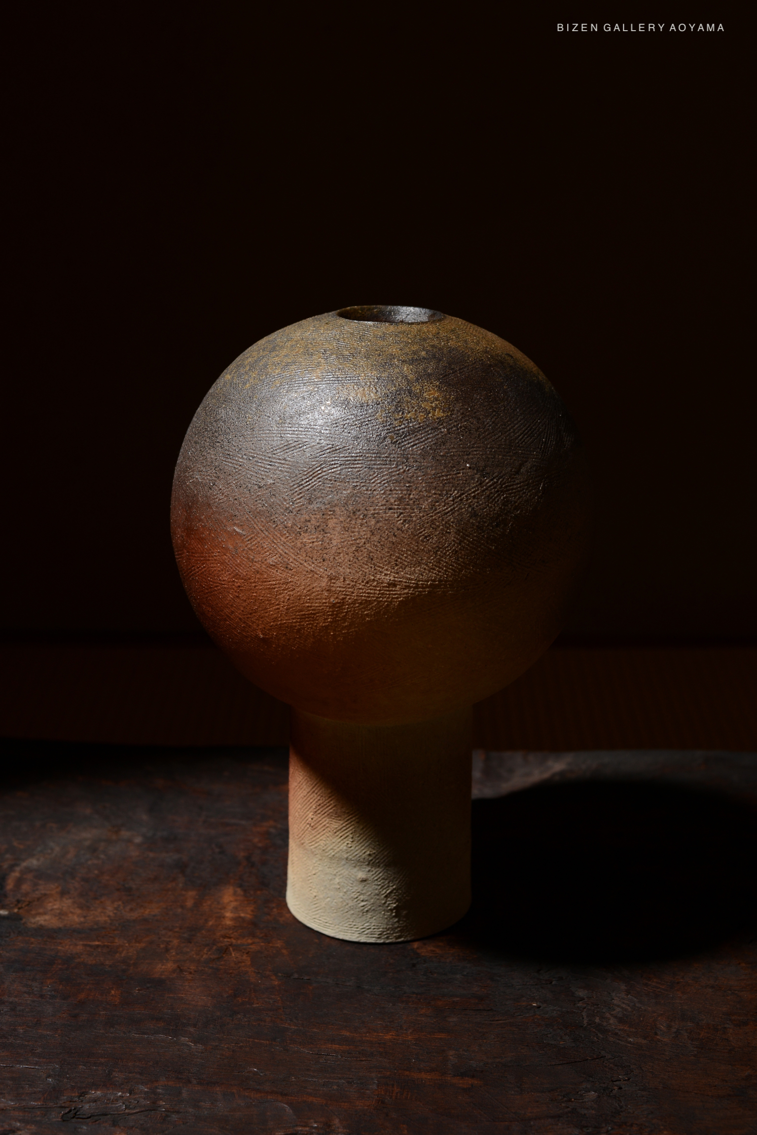 A handcrafted ceramic vase with a spherical top and cylindrical base, featuring earthy tones and a textured surface, displayed against a dark background.