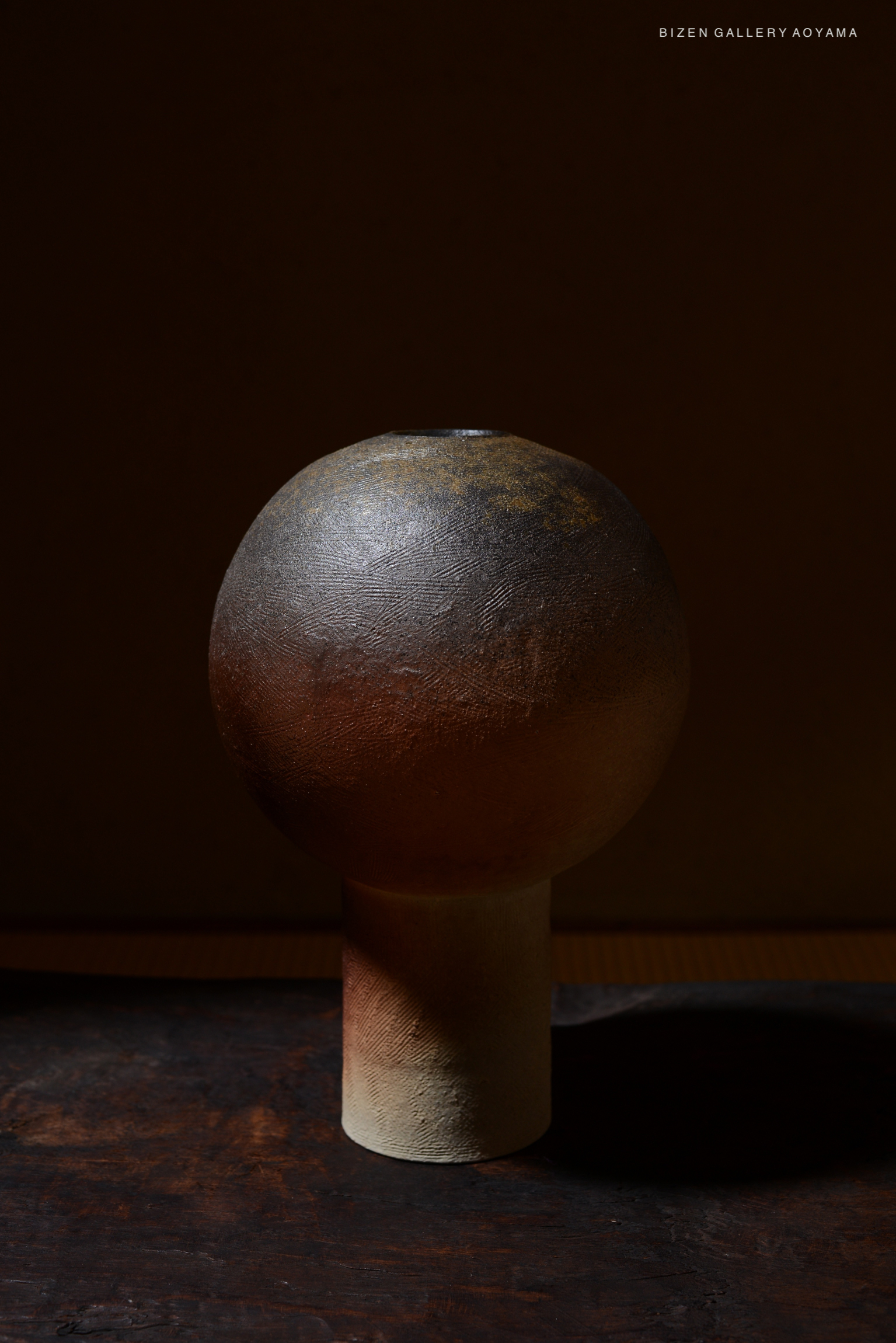A ceramic vase with a round top and a cylindrical base, showcasing a textured surface in earthy tones, photographed against a dark background.