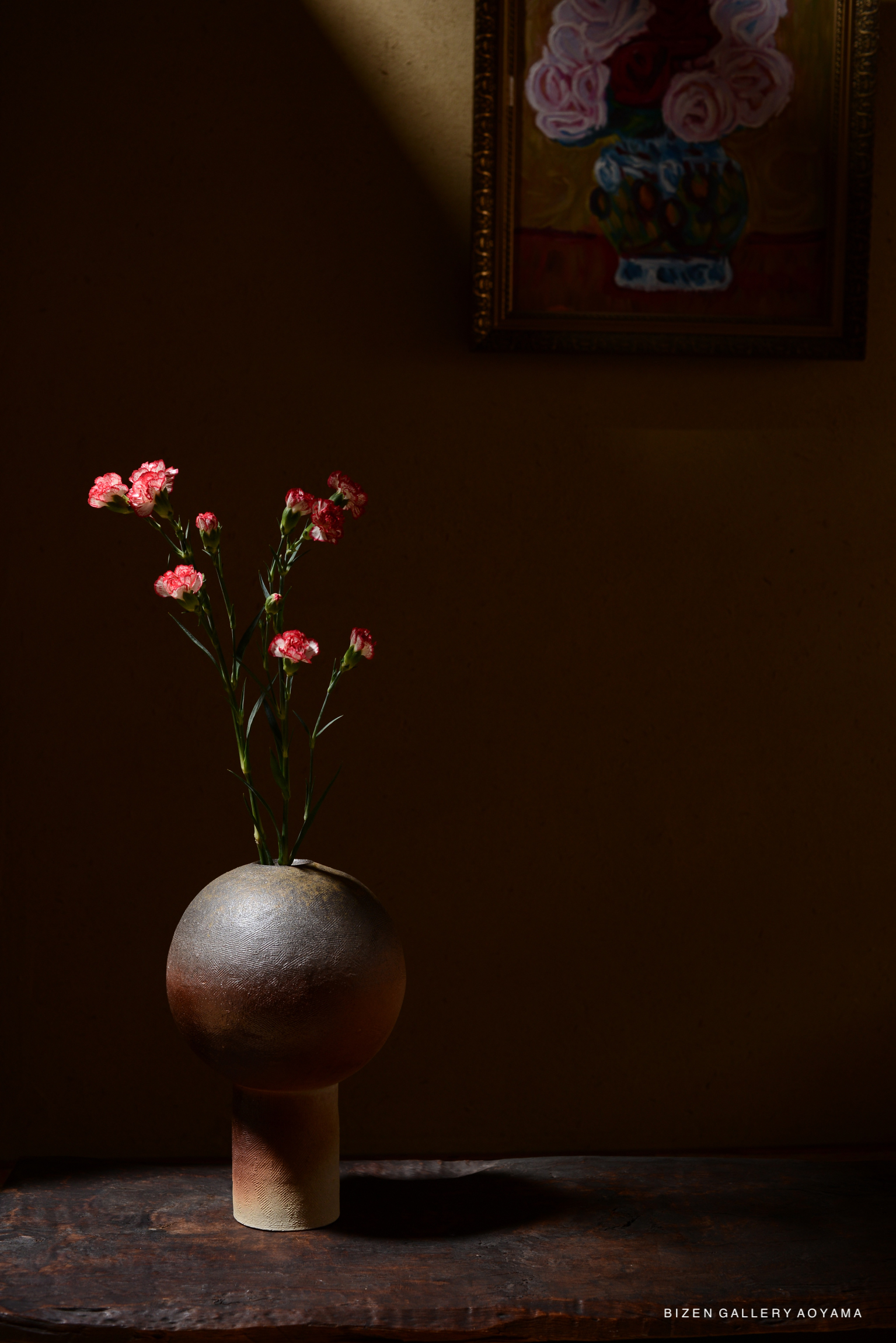 A round ceramic vase with a textured surface holding pink flowers, placed on a wooden table against a soft, shadowy background.
