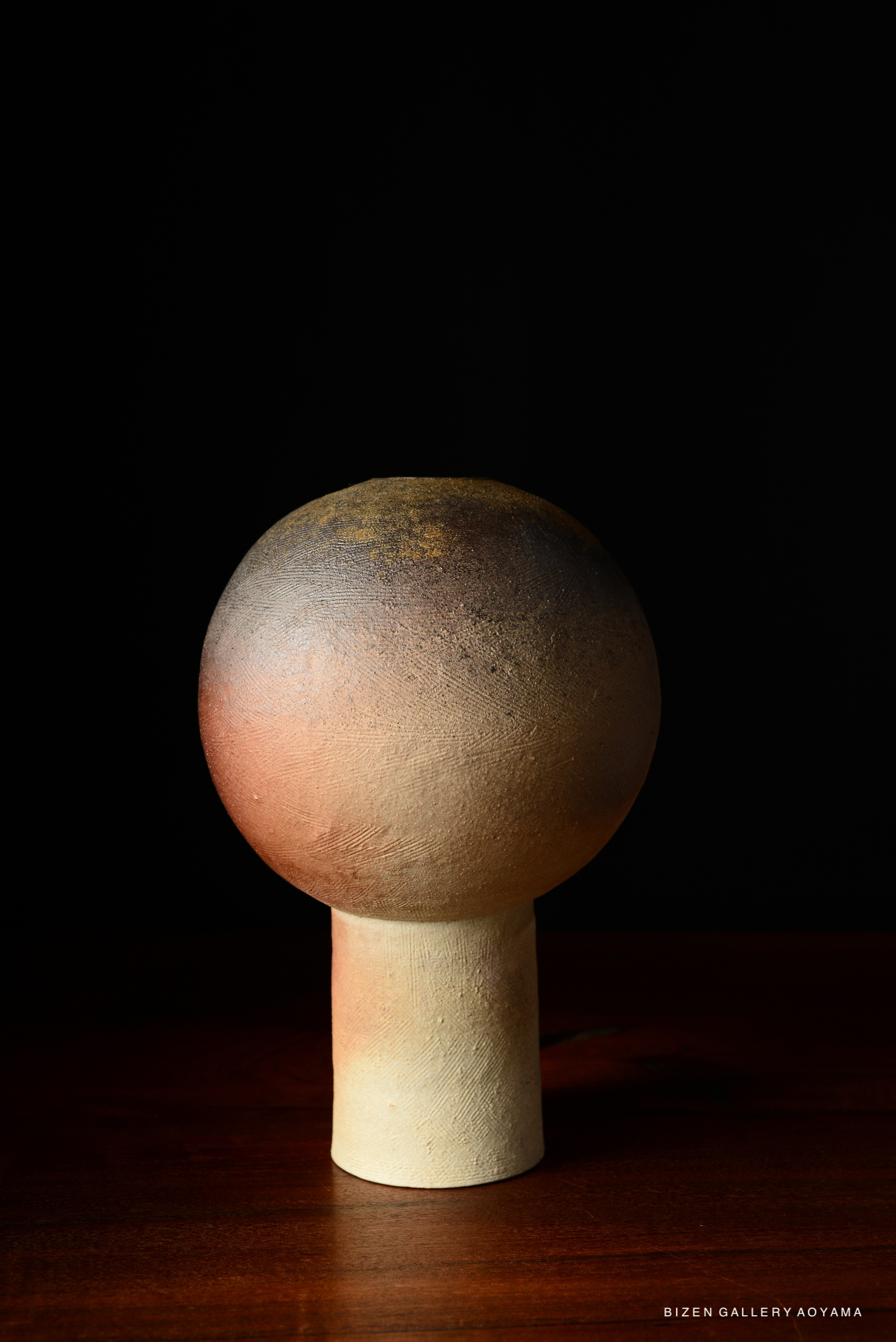 A round ceramic sculpture with a smooth, textured surface, featuring a gradient of earthy colors, displayed against a black background.