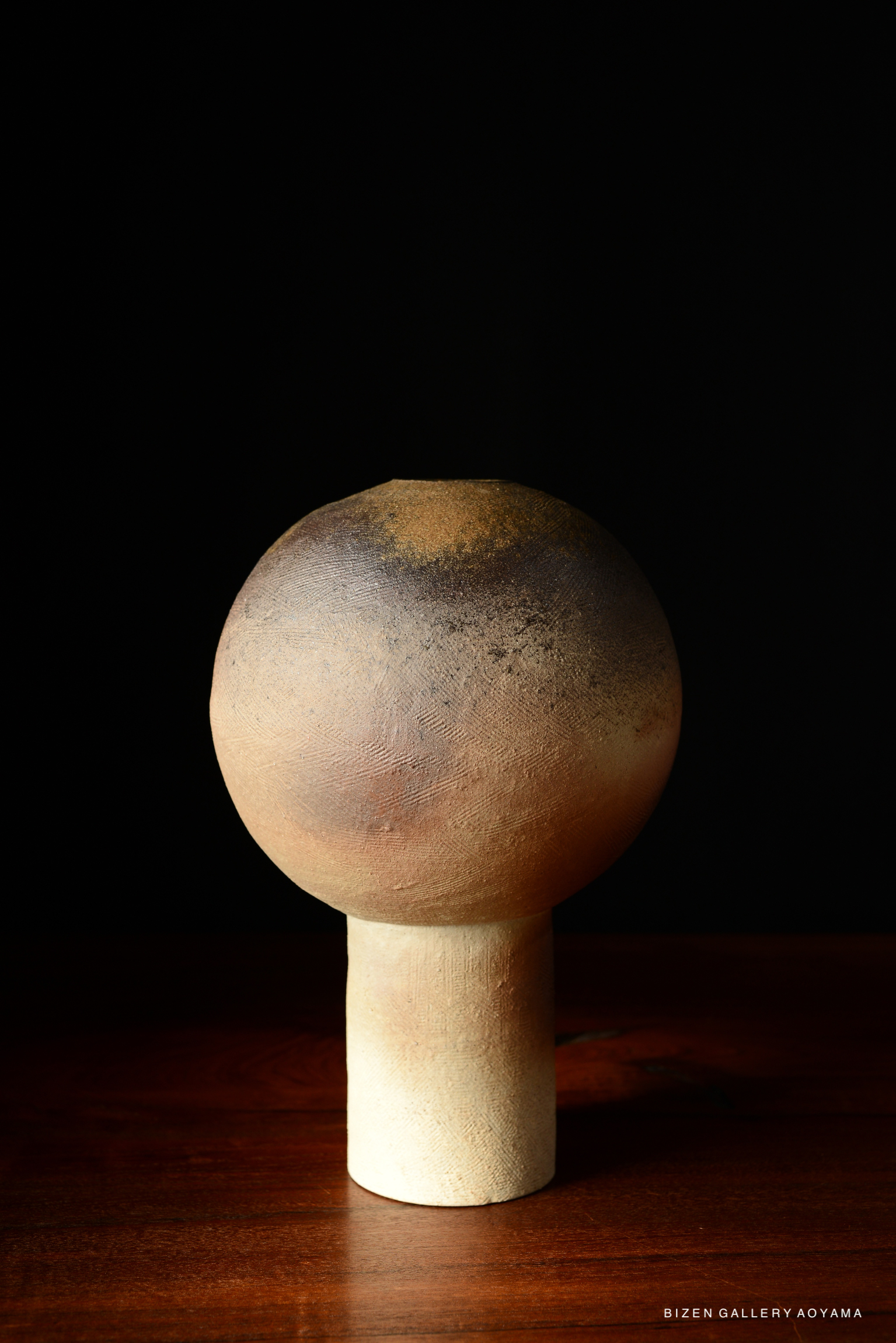 A spherical ceramic vase with a textured surface, mounted on a cylindrical base, displayed against a dark background.