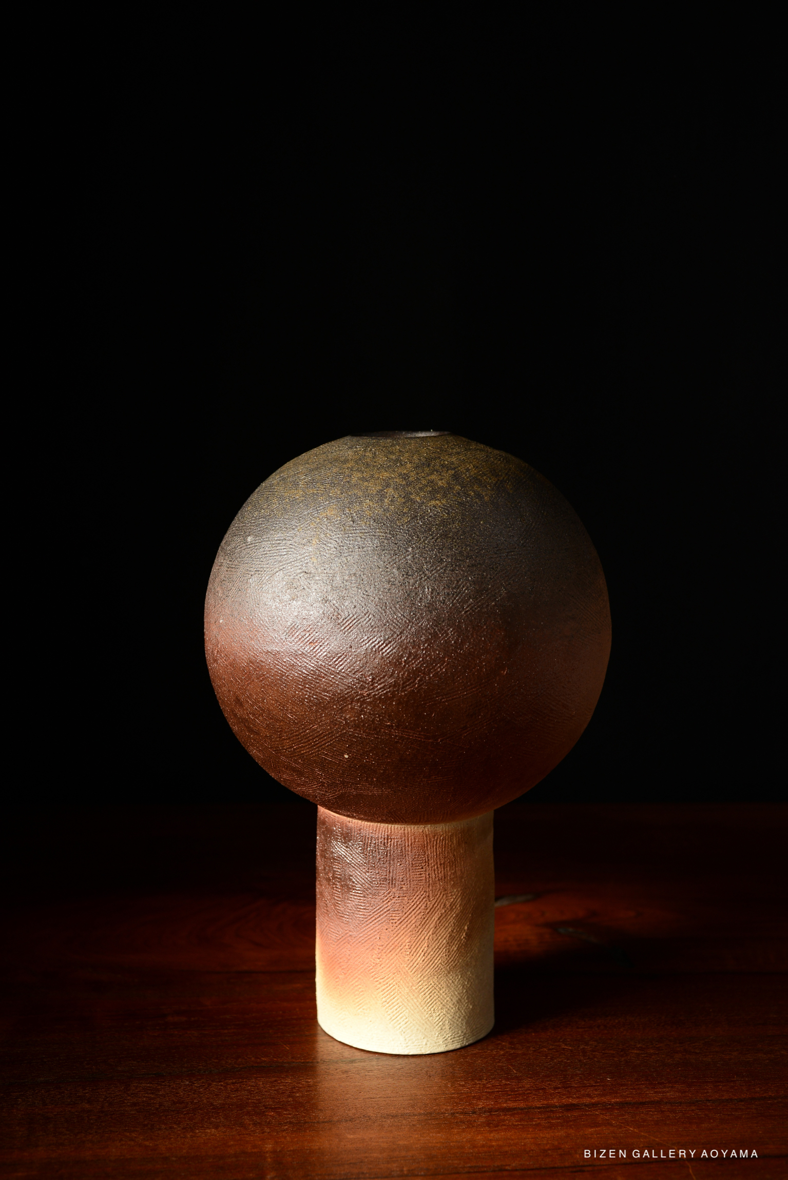 A ceramic vase with a spherical top and a cylindrical base, featuring earthy tones and a textured surface, against a dark background.