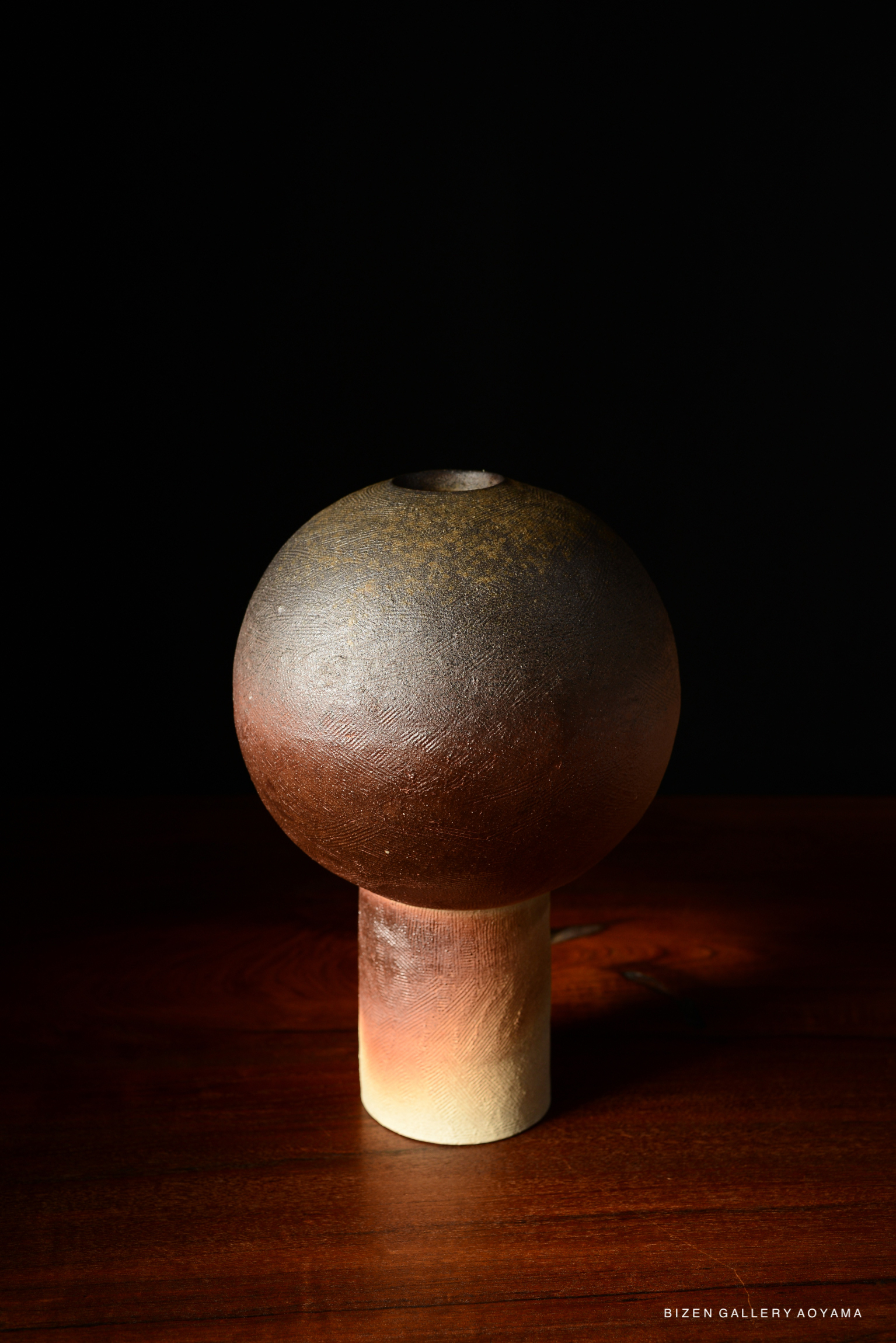A ceramic vase with a round top and cylindrical base, featuring a textured surface in earthy tones, displayed on a wooden surface against a dark background.