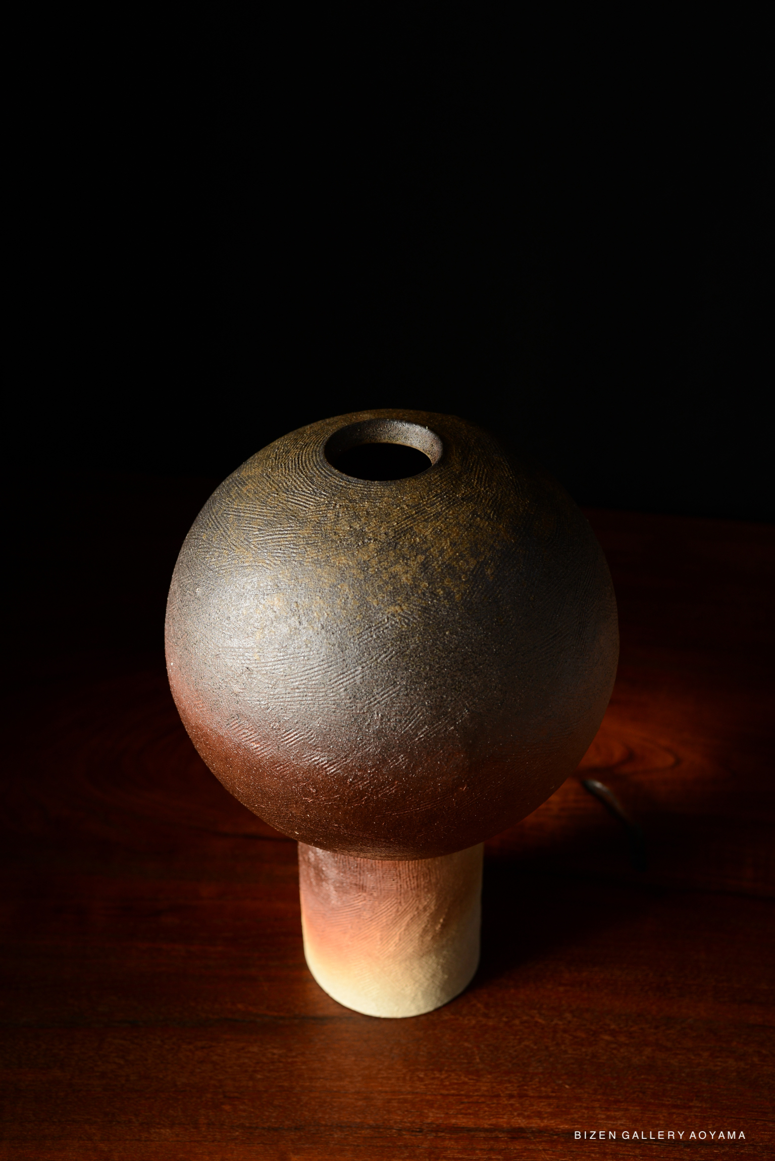 A close-up image of a round, textured ceramic vase with a narrow opening, sitting on a wooden surface against a dark background.