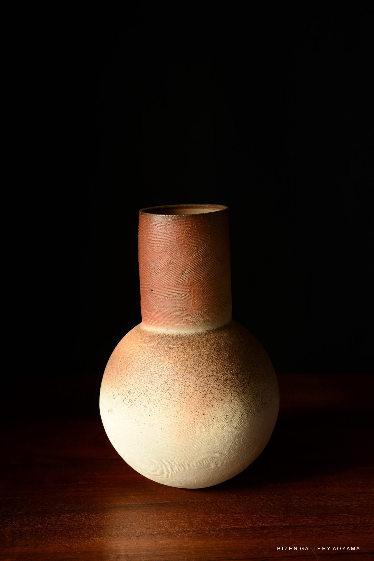 A close-up view of a handmade ceramic vase with a round body and a narrow neck, featuring a gradient of earthy tones from reddish-brown to white, displayed against a dark background.