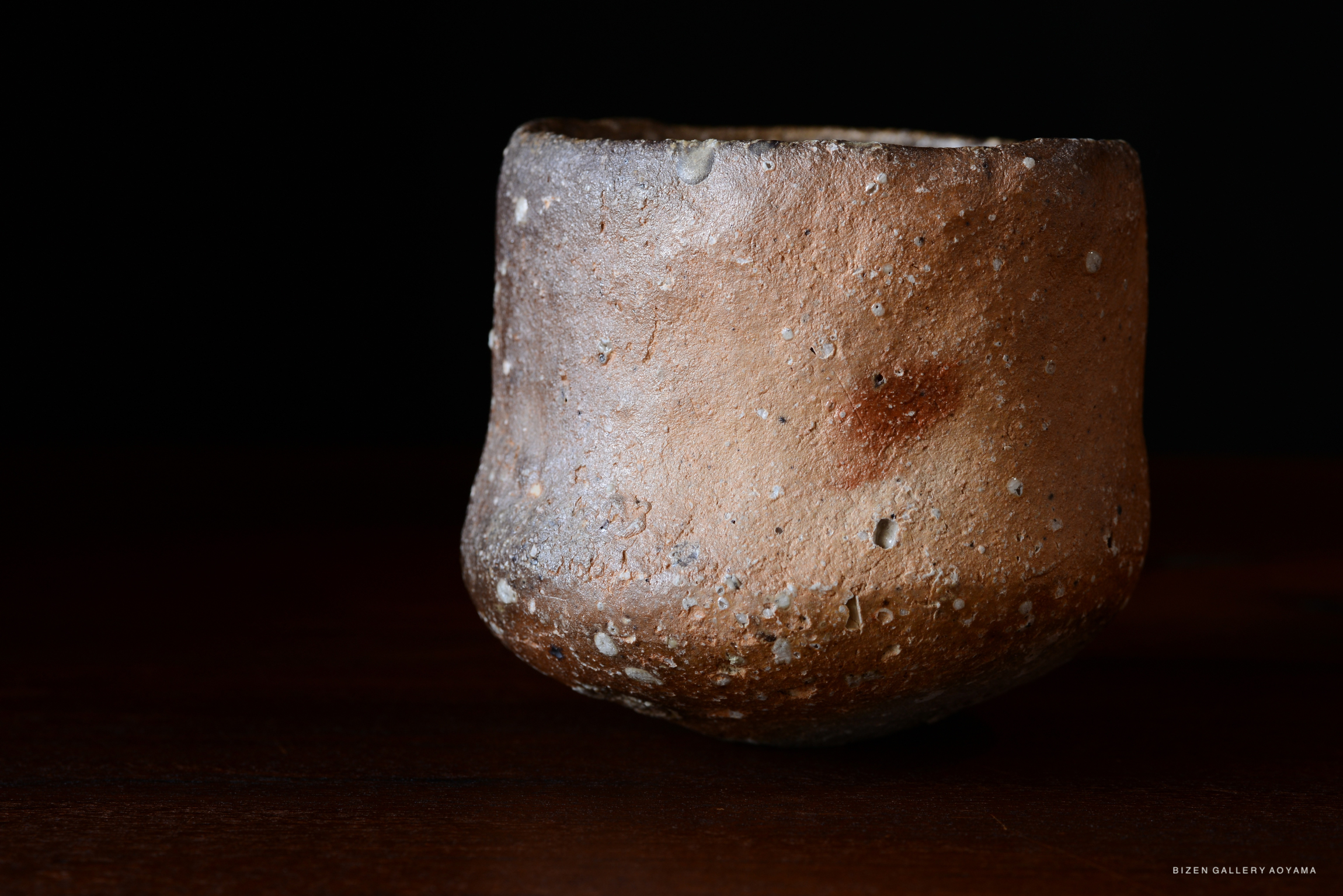 黒い背景の前に置かれた土の色の陶器の器。表面はざらざらしており、小さな粒がいくつか見える。