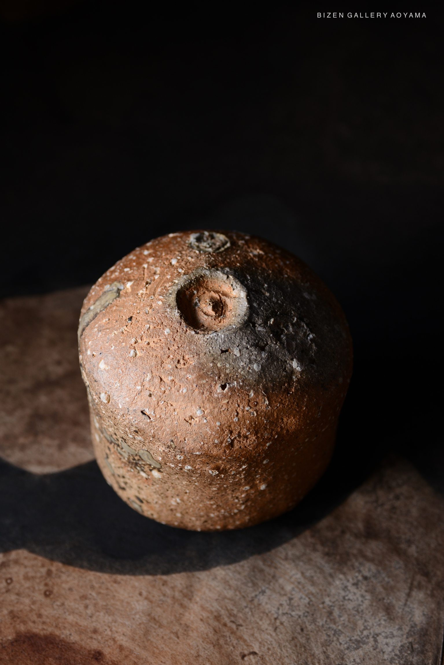 土の質感が感じられる丸い陶器の作品。暗い背景に置かれ、自然光で照らされた形状と色合いが際立つ。