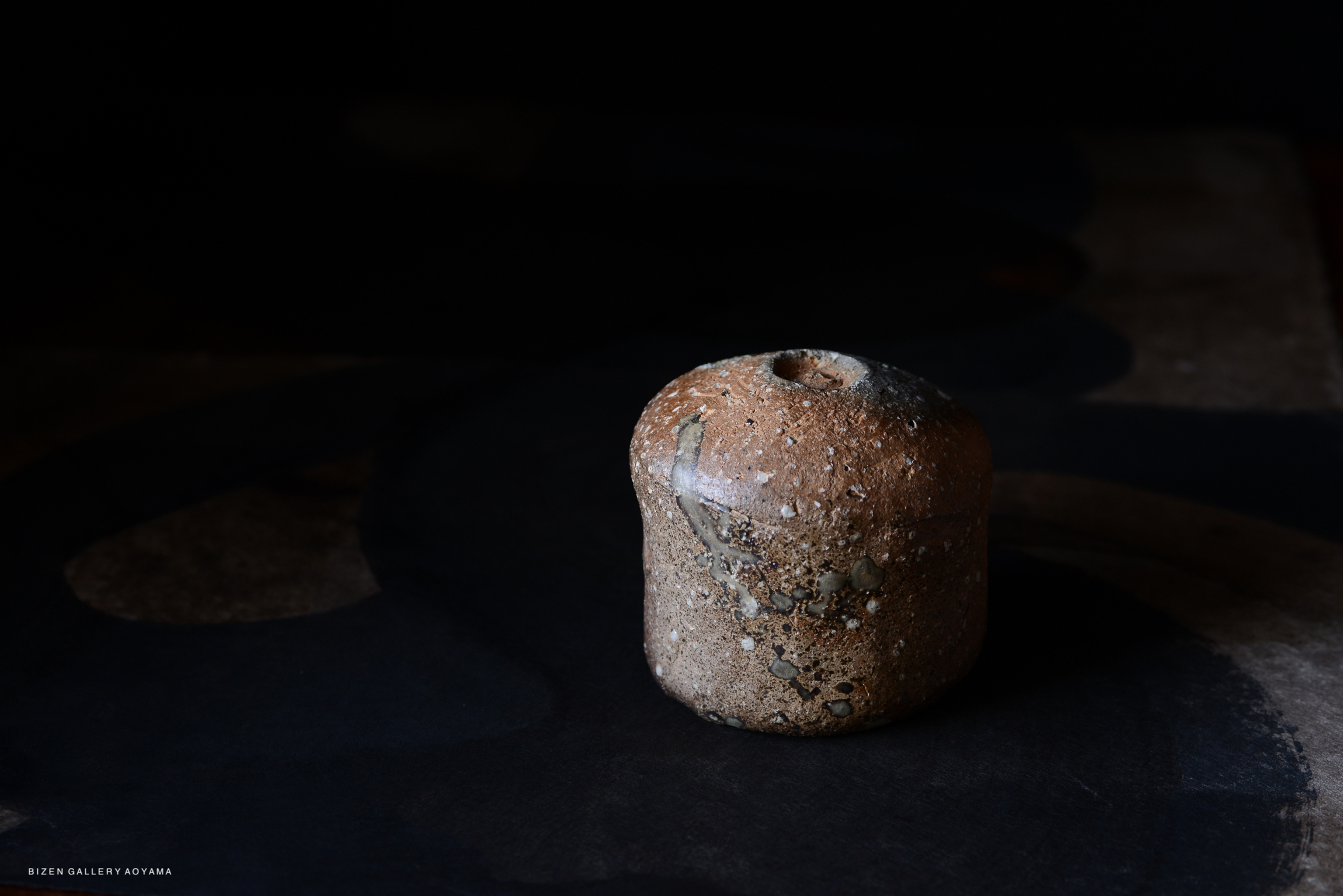 暗い背景の上に置かれた焼き物のオブジェ。陶器の表面には自然な模様があり、丸みを帯びた形状をしています。
