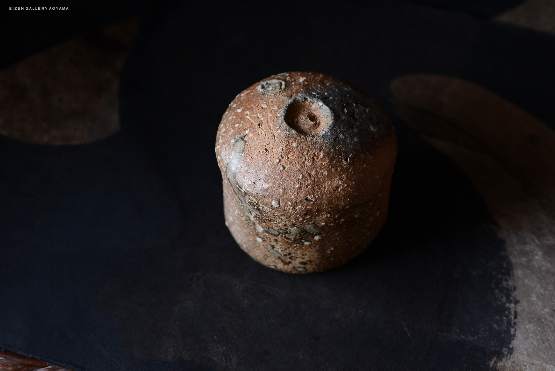 黒い背景の上に置かれた土製の容器。表面はざらざらしており、小さな穴がある。