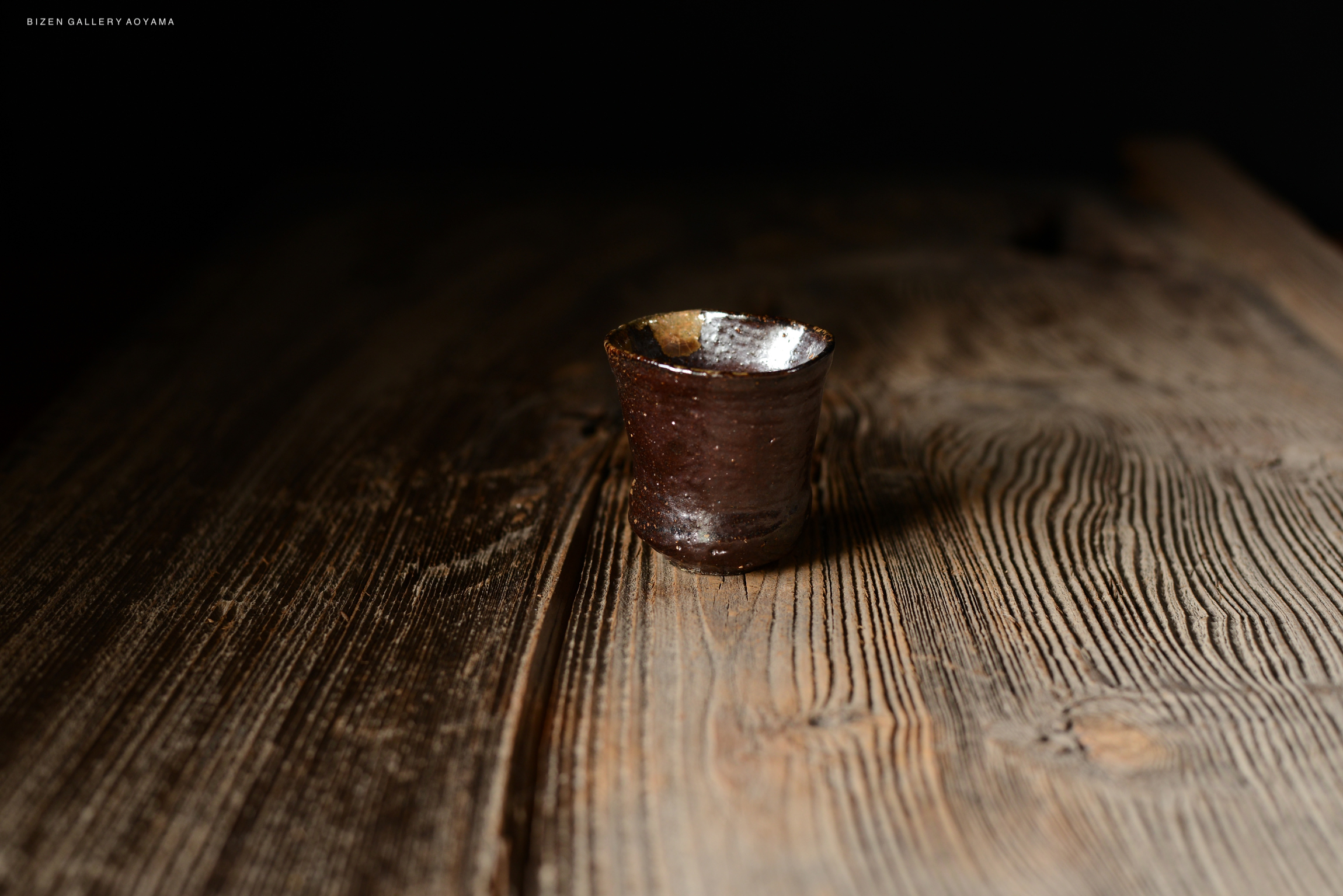 木製のテーブルの上に置かれた焼き物の杯。暗い背景で、つやのある表面が特徴。