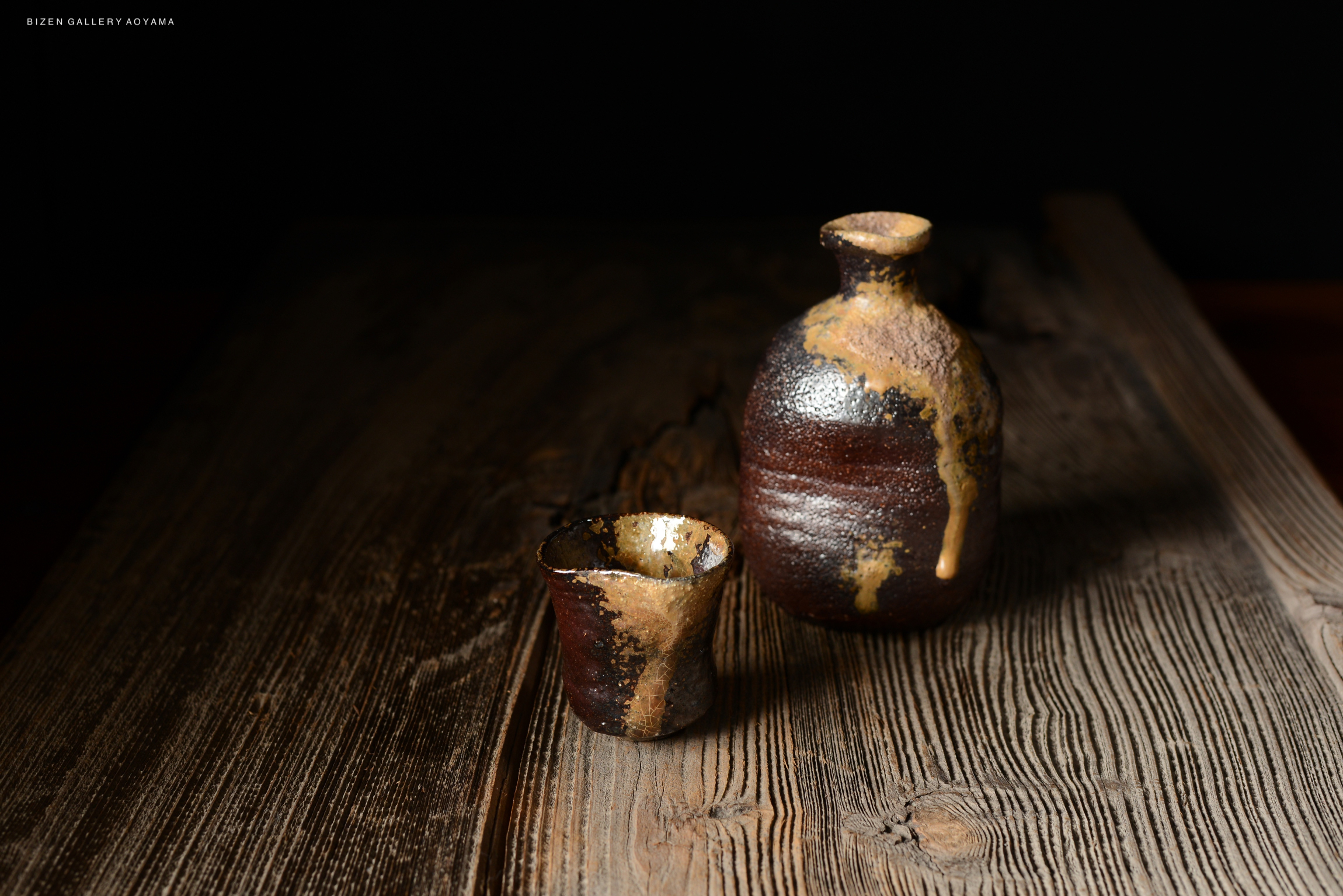 黒い背景の前に置かれた茶色の壺と小さなカップ。木製のテーブルの上にあり、土や釉薬のテクスチャーが特徴的。