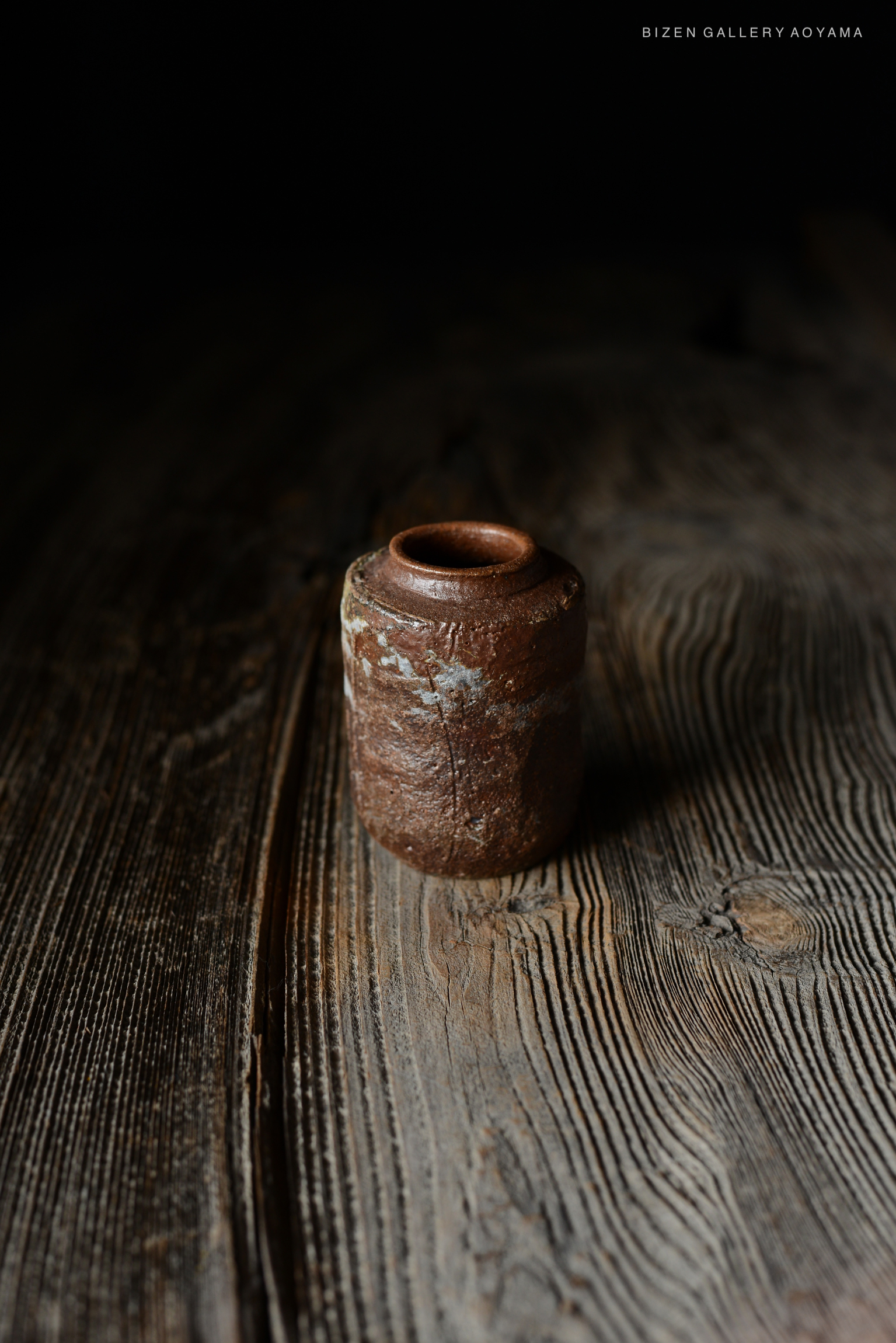 暗い背景に置かれたビゼン焼きの小さな壺が、木のテーブルの上で強調されている