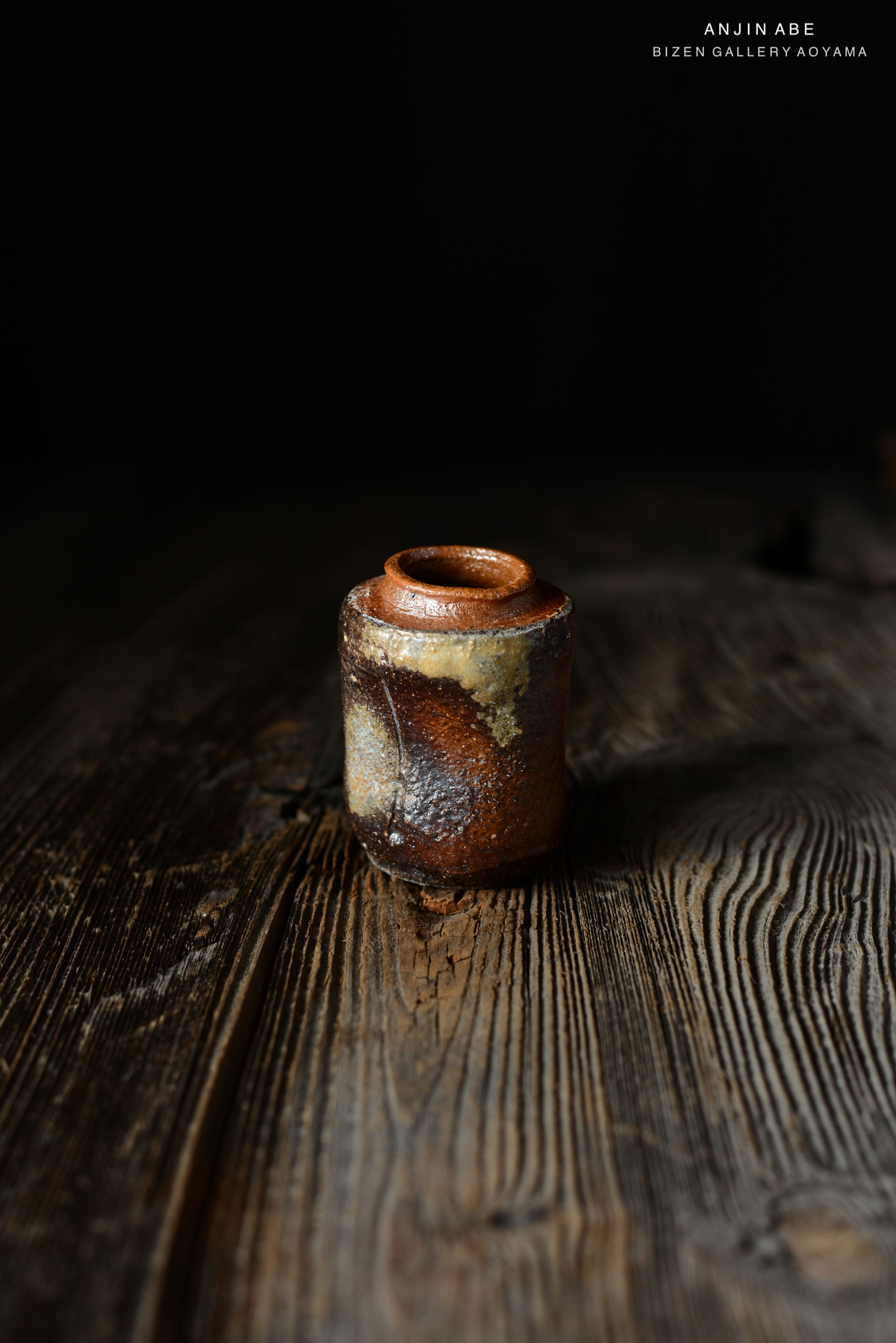 黒い背景の前に置かれた、独特の釉薬が施されたビゼン焼きの小さな壺。木のテーブルの上に置かれている。
