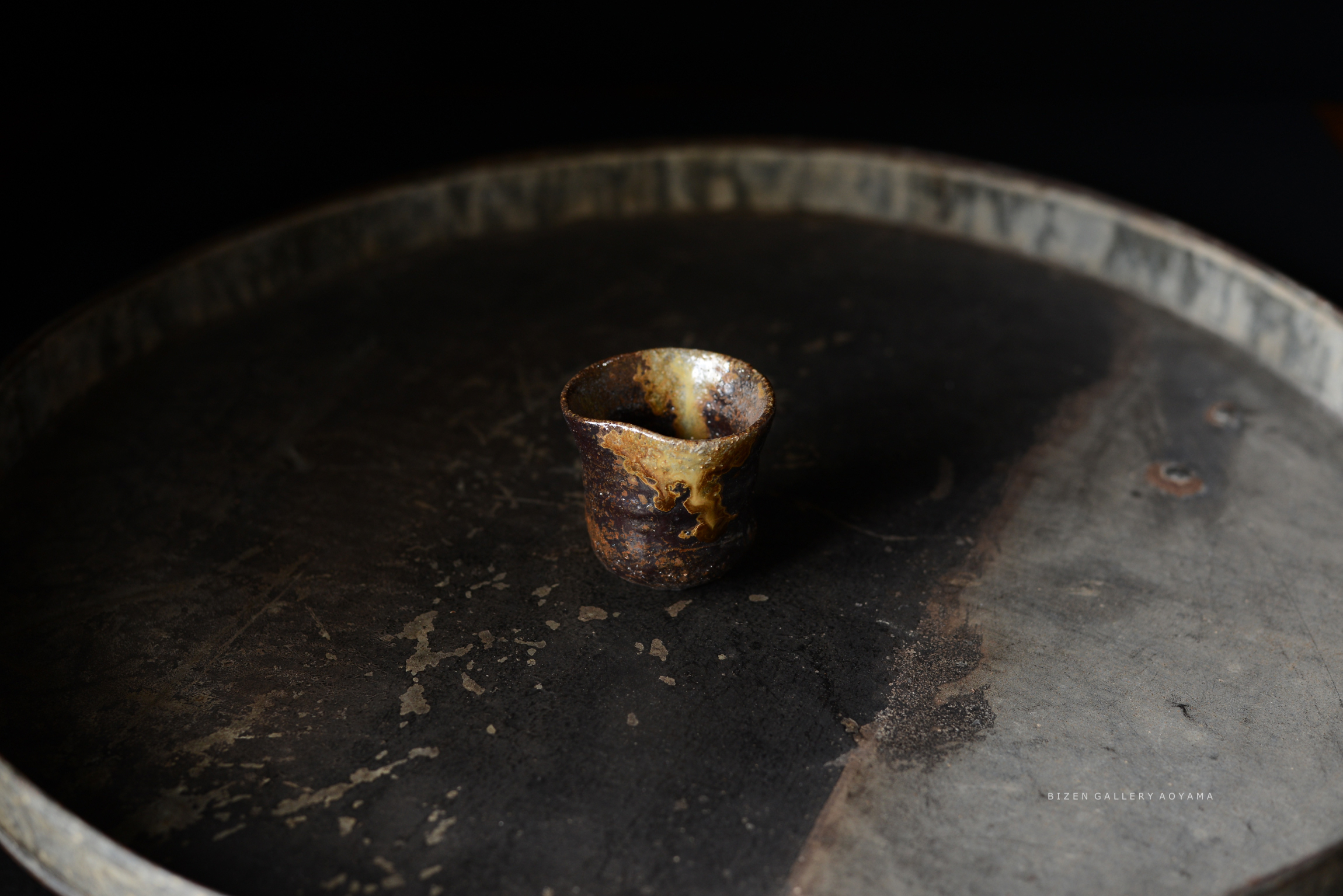 黒い背景の上に置かれた、独特な形状の陶器のカップ。カップは赤褐色で、表面に釉薬の流れが見られる。