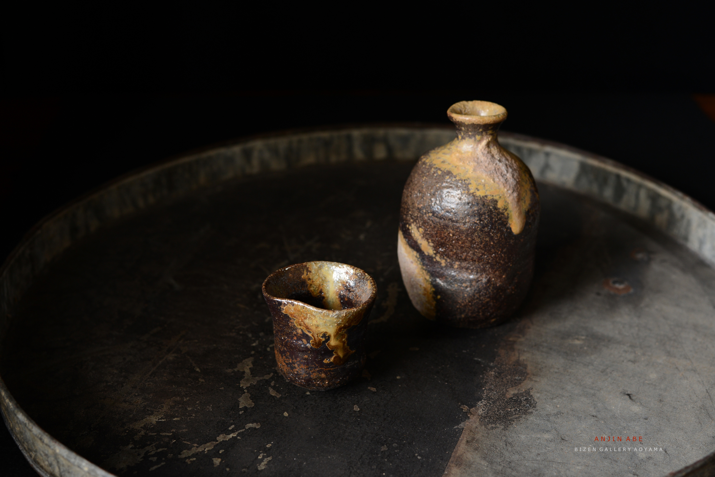 黒いトレーの上に置かれた土器の壺と杯。壺は丸みを帯びた形状で、独特の釉薬の模様が見られる。杯は小ぶりで、同様の色彩を持っている。