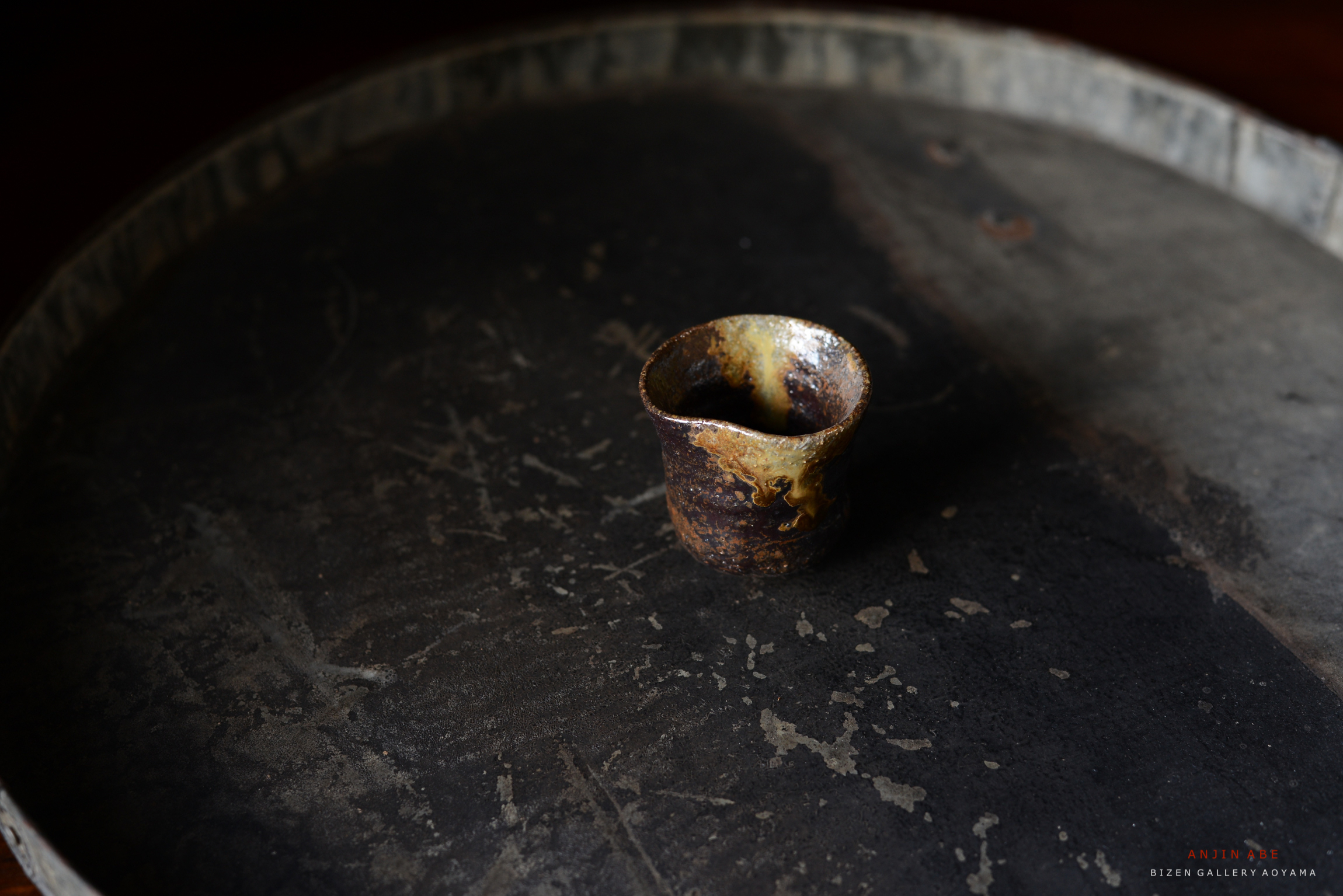 黒い皿の上に置かれた土製の小さなカップ。カップは茶色と金色が混ざった独特の釉薬で装飾されている。