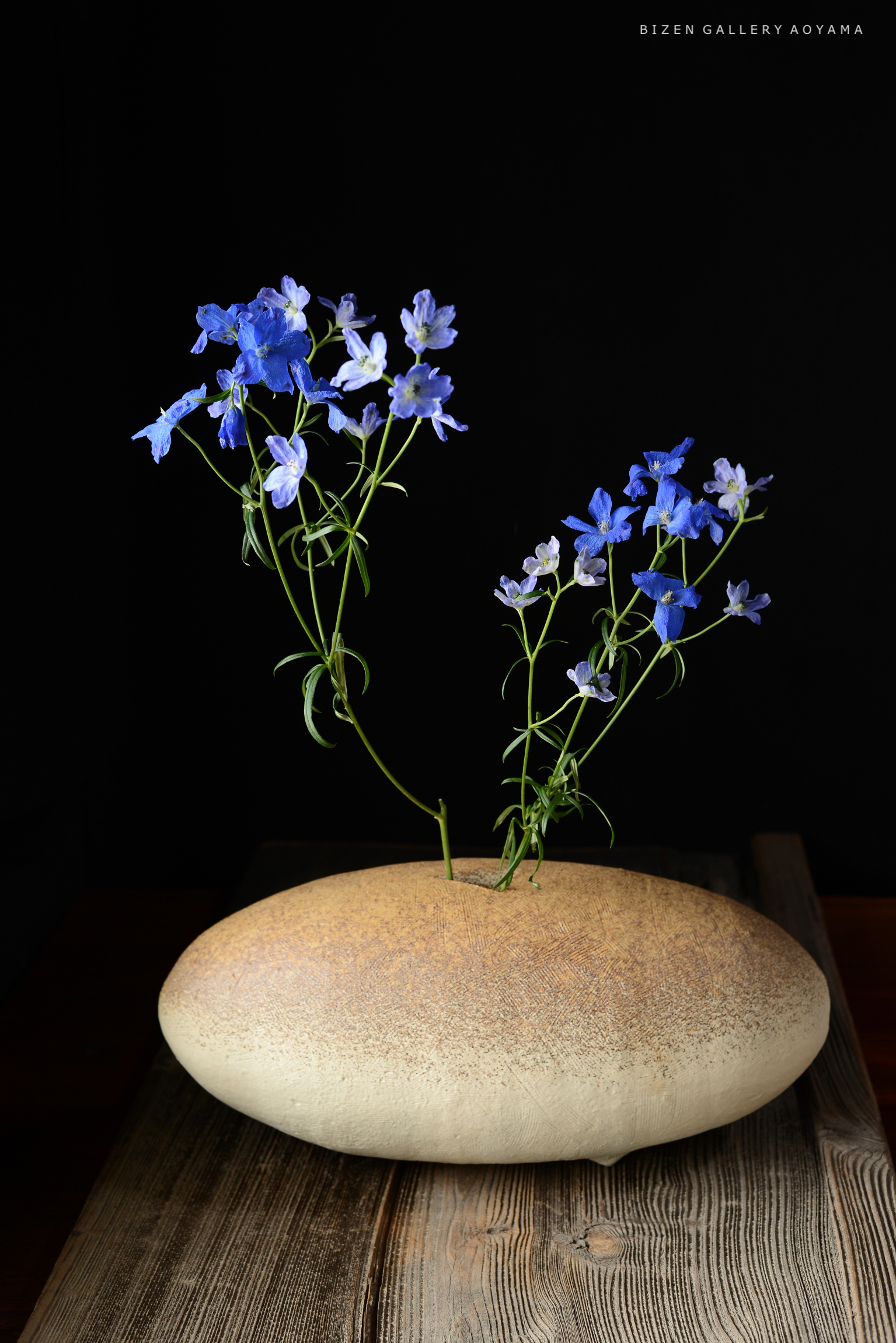 青い花が特徴的なフラワーベースが木製のテーブルの上に置かれている。背景は黒色。