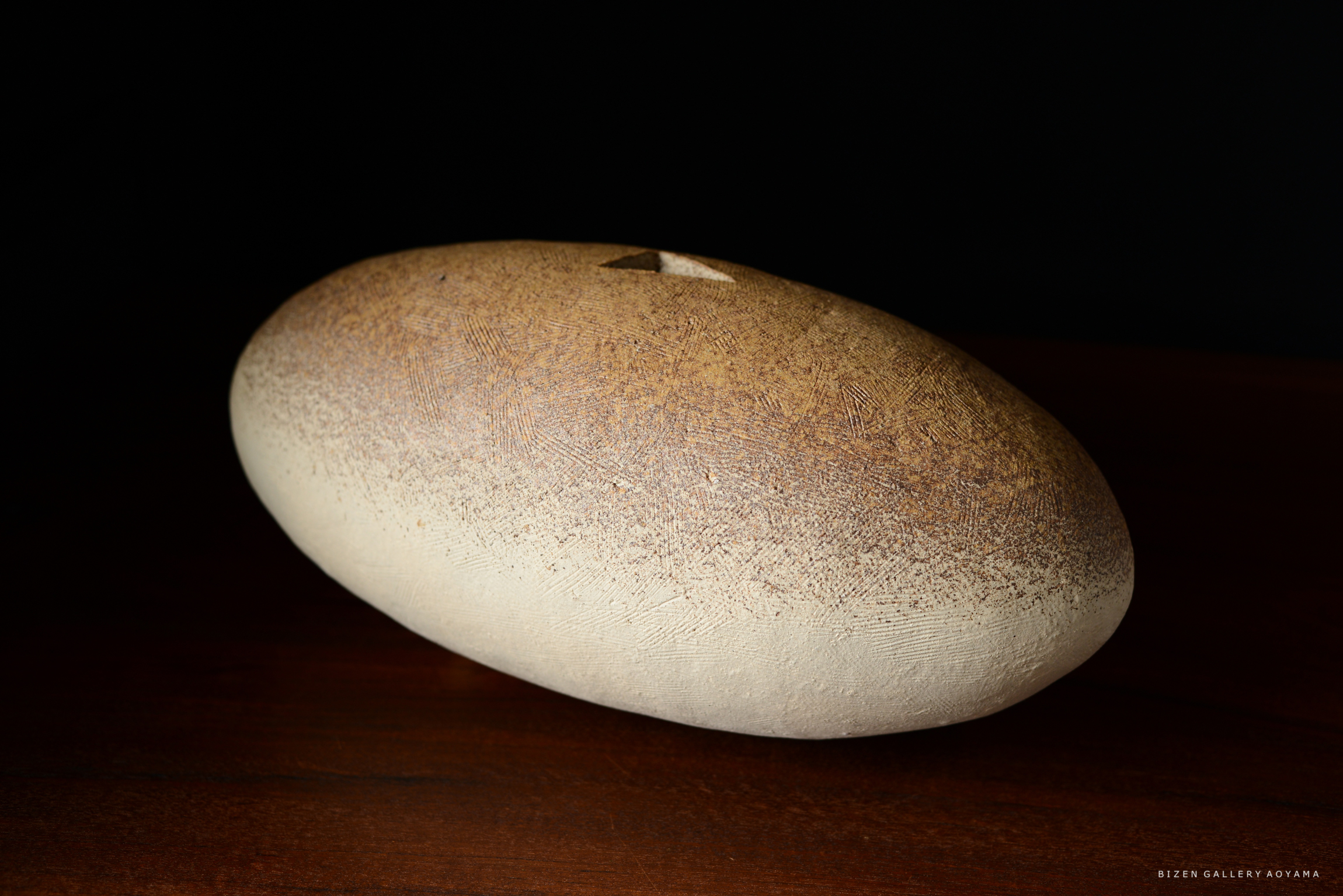 円形の陶器のオブジェ、滑らかな表面と茶色のグラデーションが特徴。暗い背景と木のテーブルに置かれている。