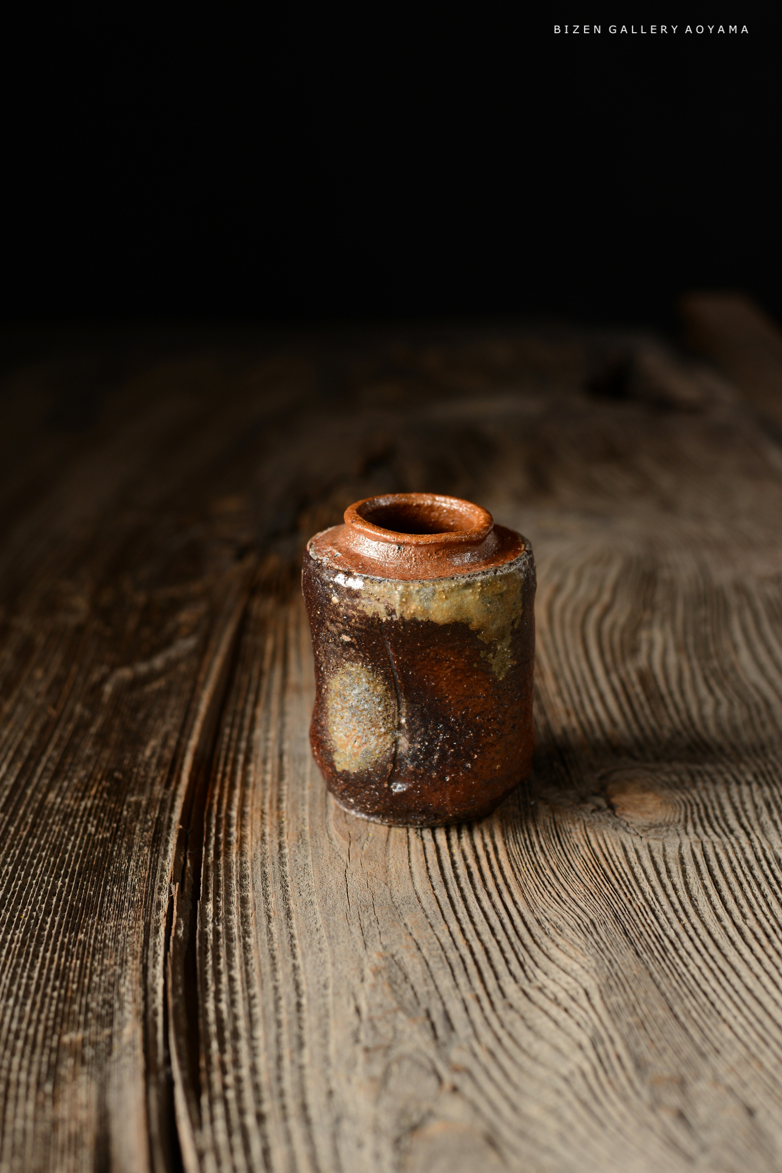 焼き物の小さな瓶が木のテーブルの上に置かれています。瓶は釉薬の異なる色合いを持ち、自然な質感が感じられます。