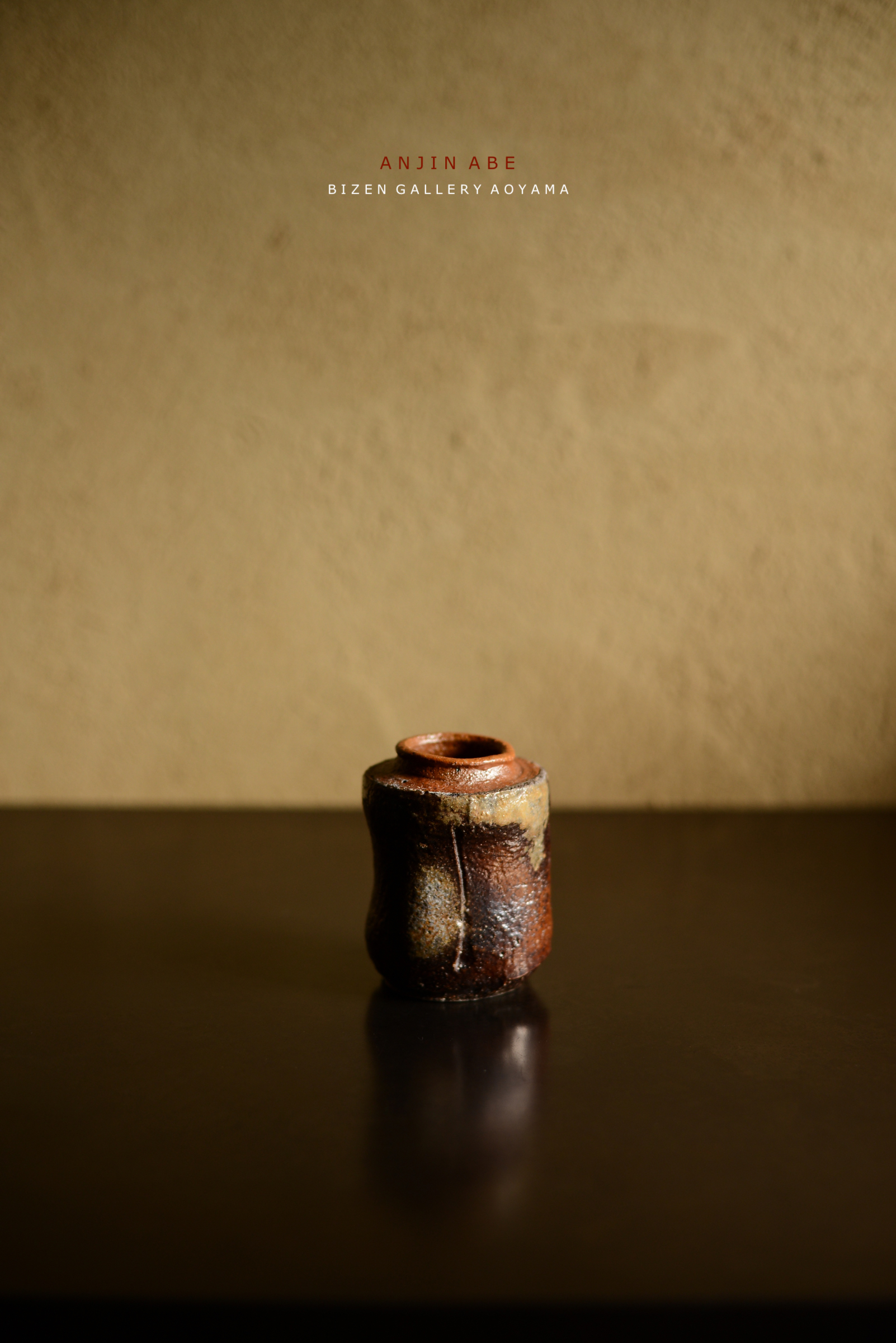 濃い色合いの備前焼の小さな壺がテーブルの上に置かれている。背景は淡い色合いの壁で、壺の前面には釉薬のかすかな模様が見える。