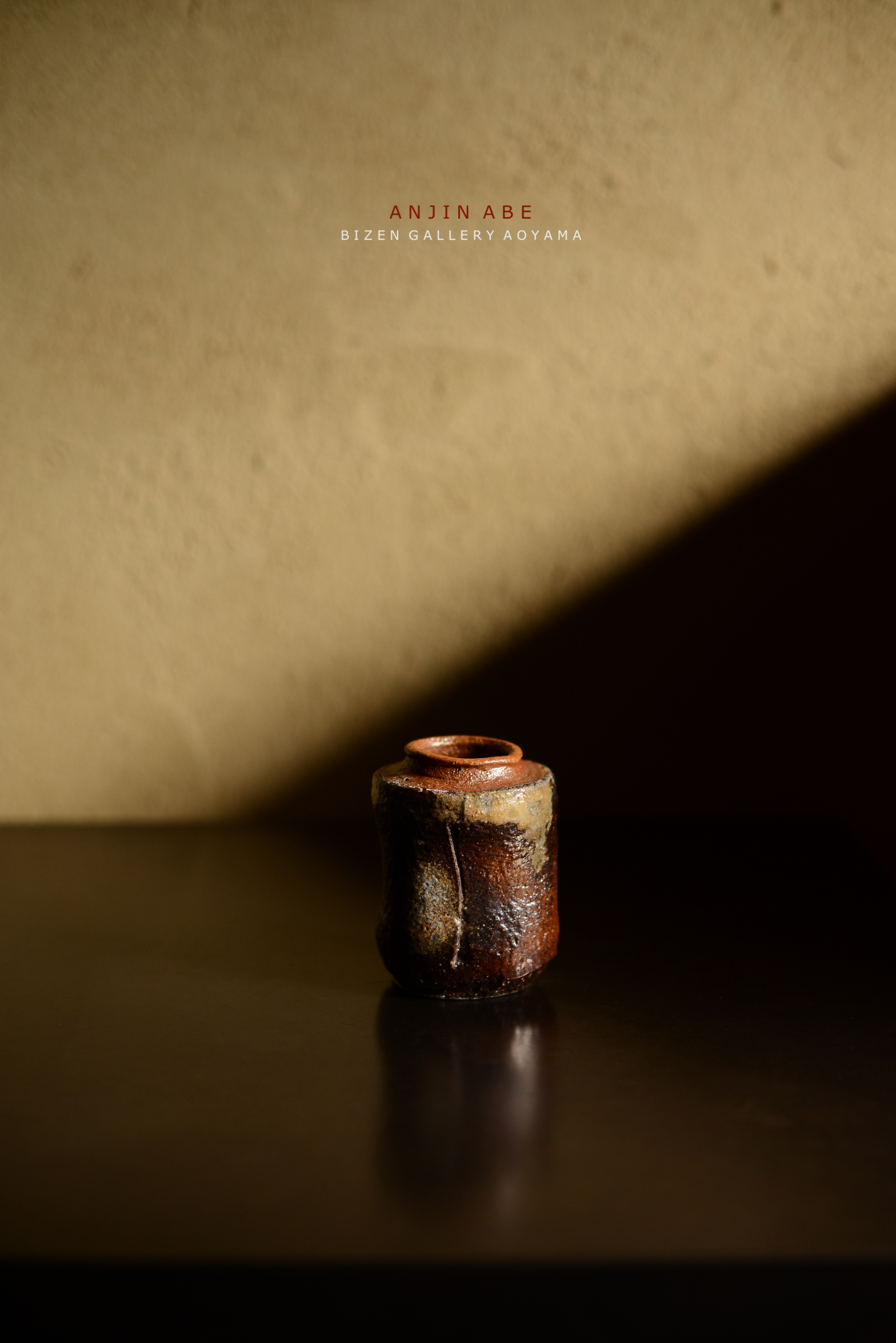 暗い背景の上に置かれた陶器の壺。壺は有機的な形状を持ち、茶色と黒の色合いが見られる。光が当たり、陶器の質感が際立っている。